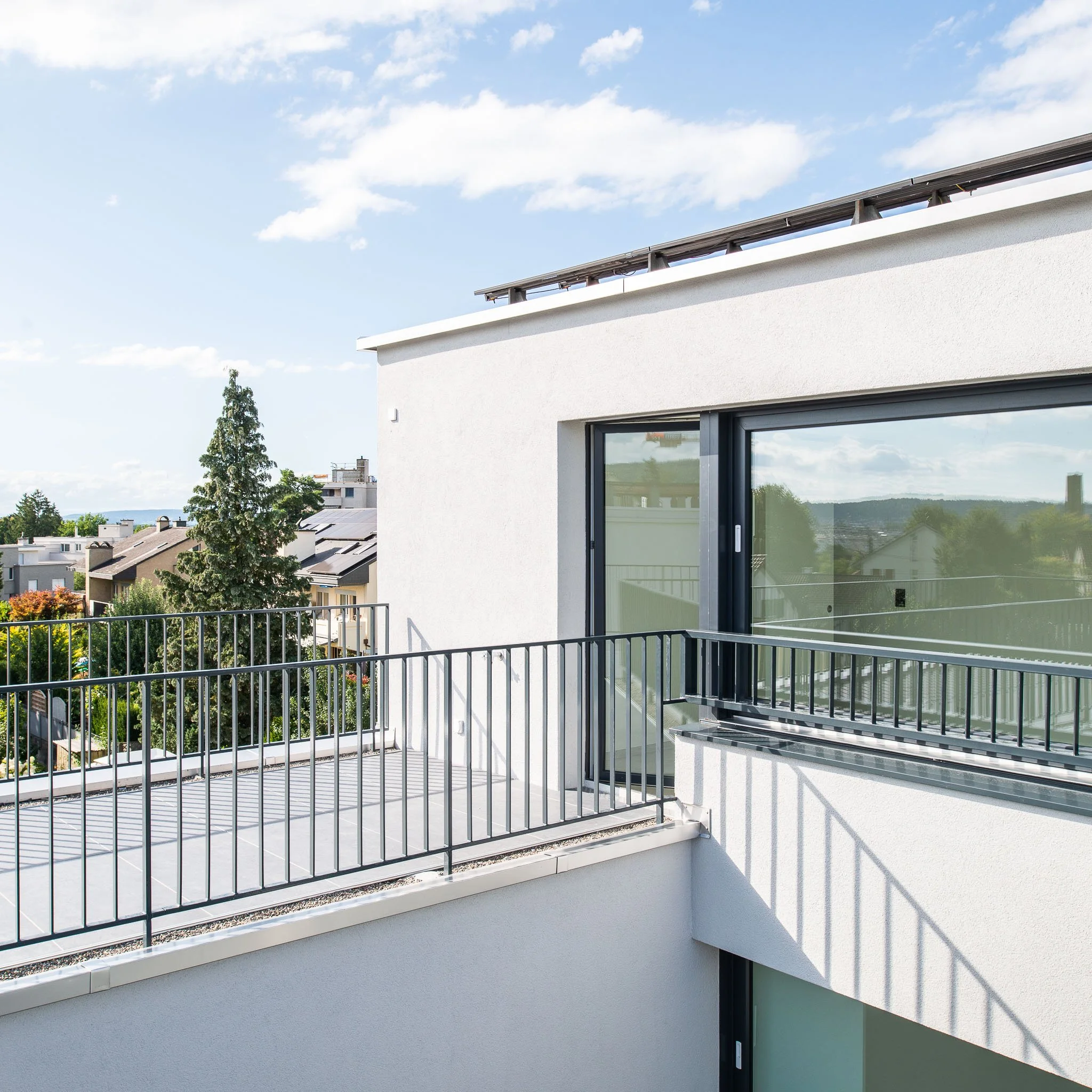 Terrasse eines modernen weissen Hauses mit Glastür und Metallgeländer, im Hintergrund ein blauer Himmel mit Wolken und grüne Bäume.