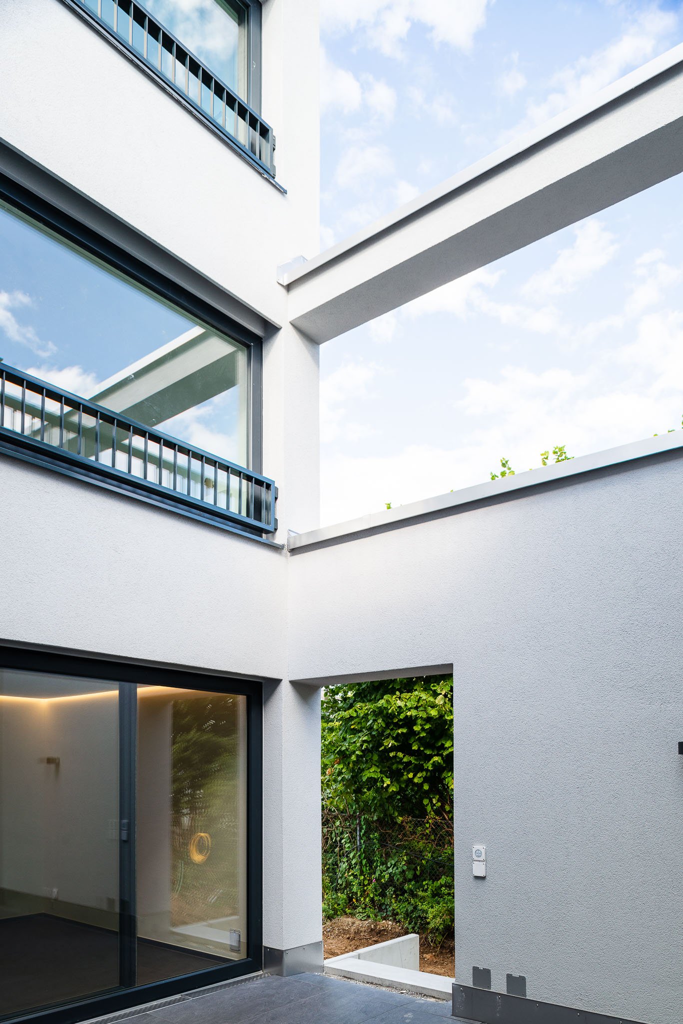Moderne weisse Hausfassade mit grossen Fenstern, Balkon mit Metallgeländer und Garten mit Bäumen im Hintergrund bei sonnigem Himmel