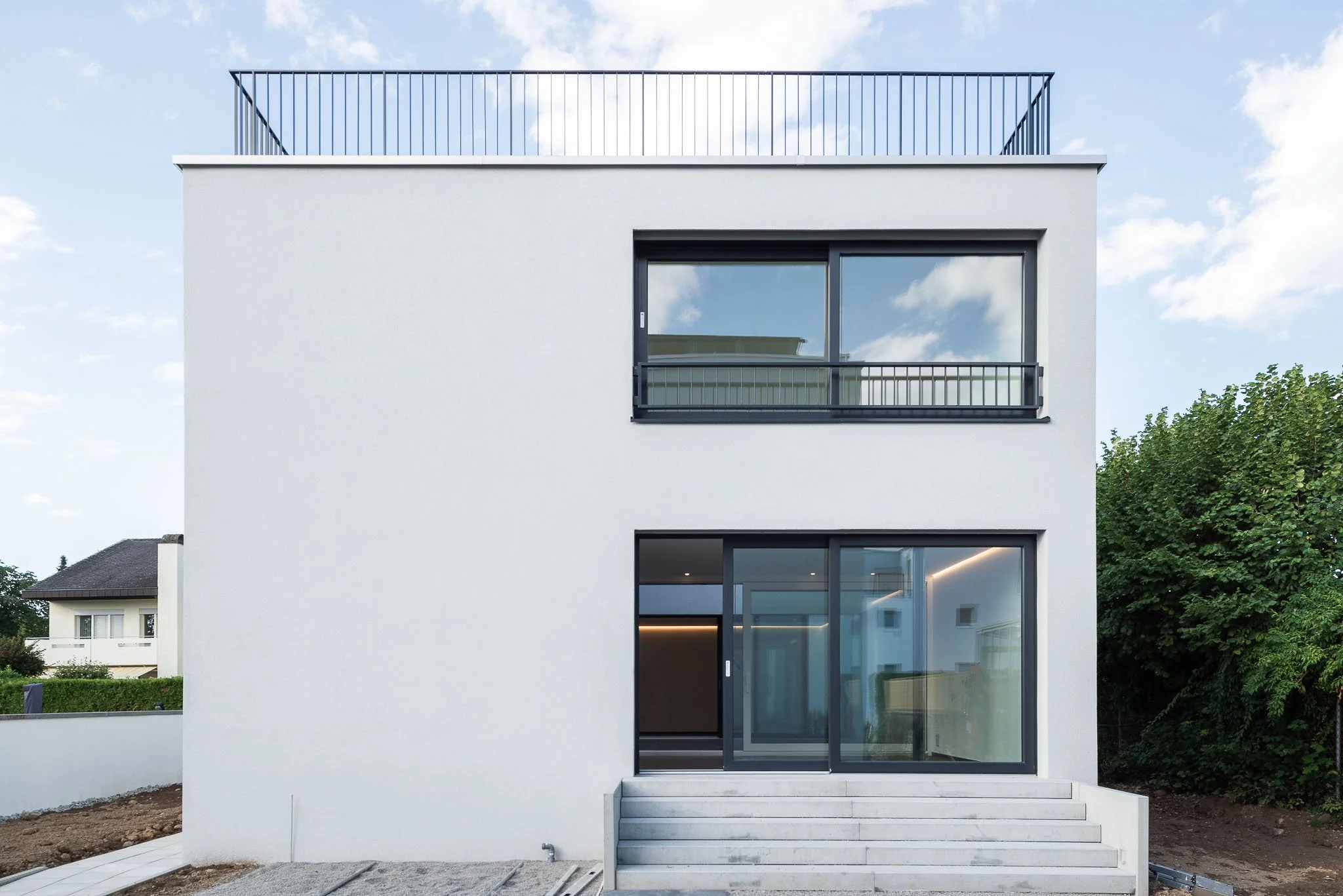 Moderne weisse Einfamilienhausfassade mit Glasfront, Balkonen und Steintreppe vor Eingang, umgeben von grünem Garten, am Tag mit blauem Himmel und Wolken