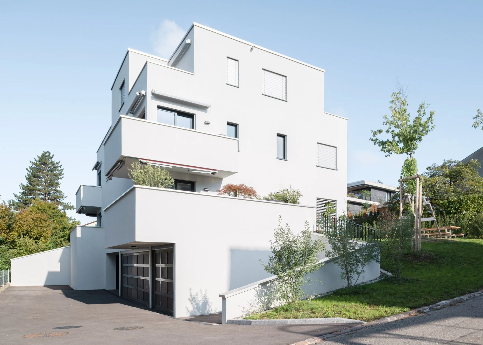 Modernes weisses Mehrfamilienhaus mit mehreren Balkonen und grossen Fenstern, umgeben von Grünflächen und Bäumen, bei Tageslicht.
