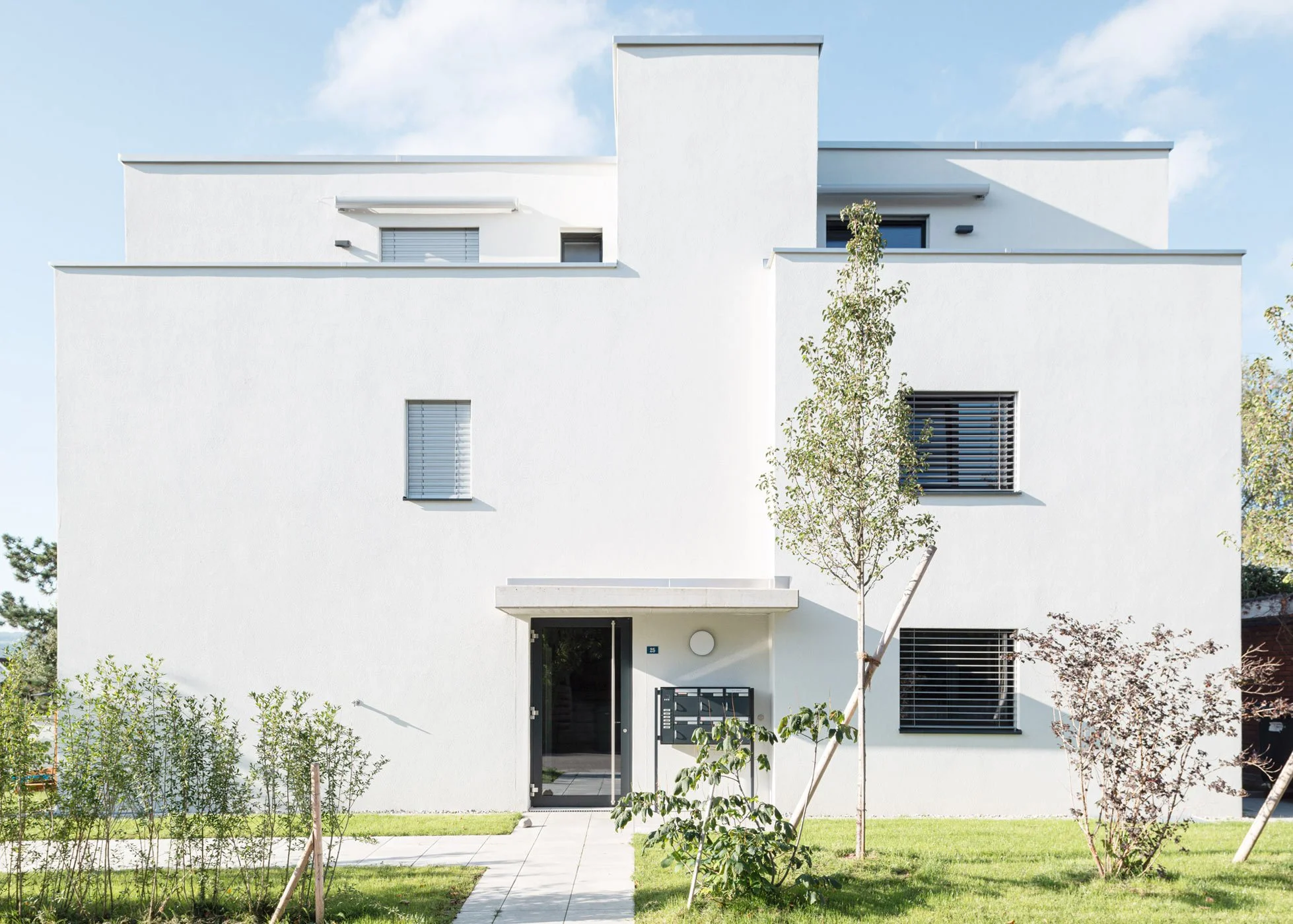 Modernes, weisses Mehrfamilienhaus mit mehreren Stockwerken, Rechtecken und Balkonen. Fenster mit Rollläden, ein Eingang mit Haustür und Briefkasten. Garten mit Bäumen und Rasen vor dem Haus.