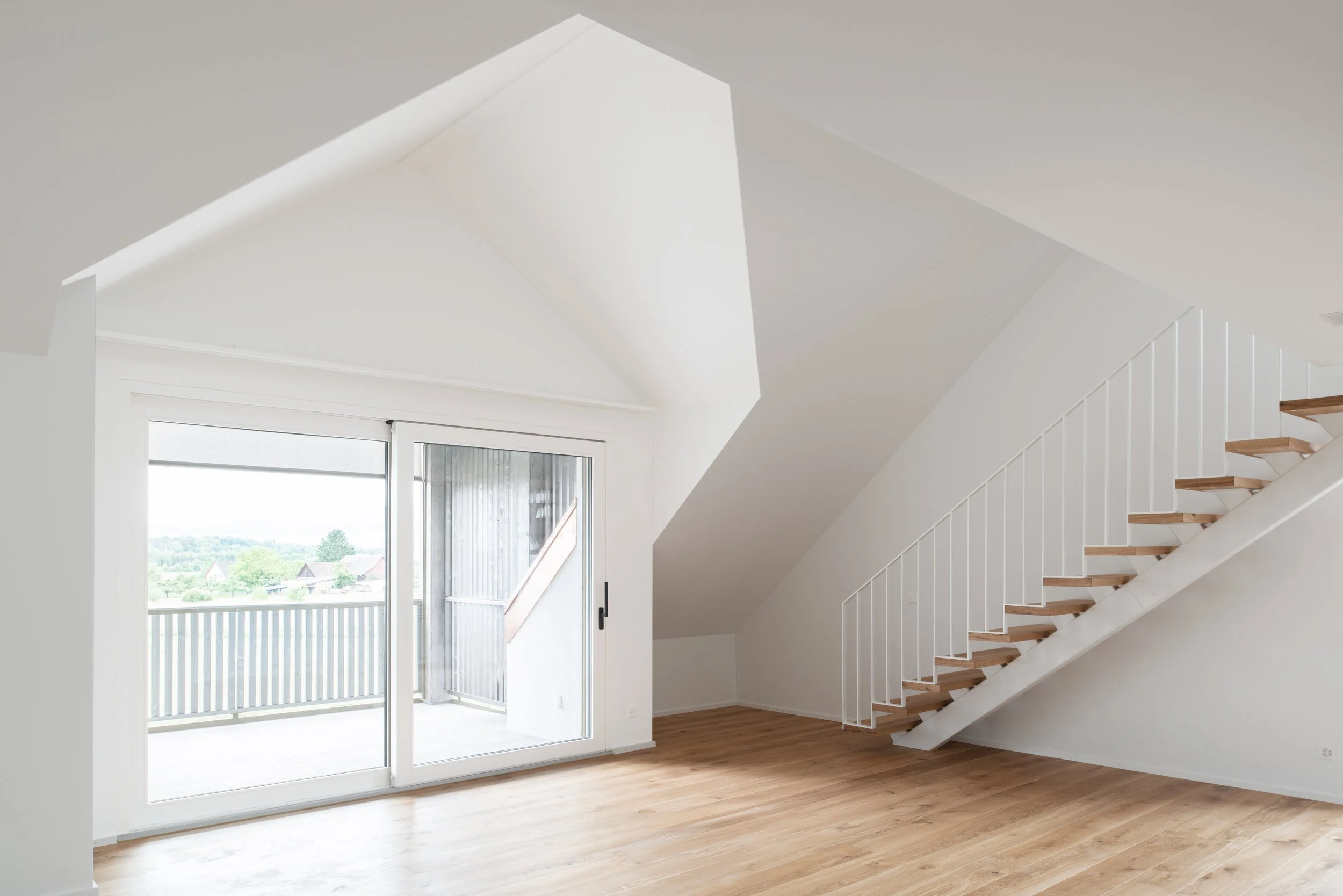 Innenraum mit weisser Wand, Holzboden und Treppe, grosses Fenster mit Balkon vor Blick auf eine grüne Landschaft.