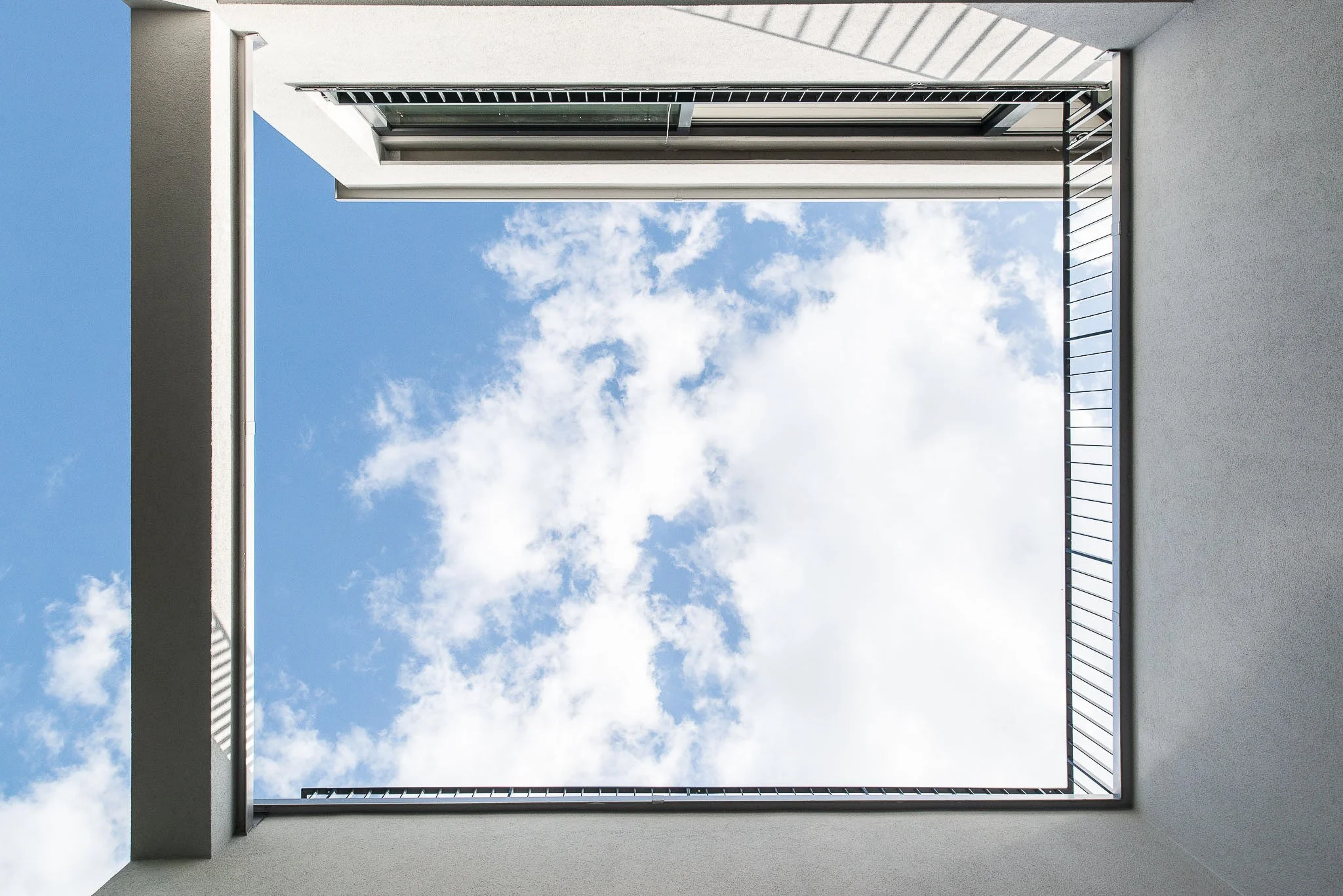 Blick nach oben durch einen offenen Innenhof zu einem blauen Himmel mit Wolken, umgeben von weissen Gebäudewänden und einem Balkongeländer.