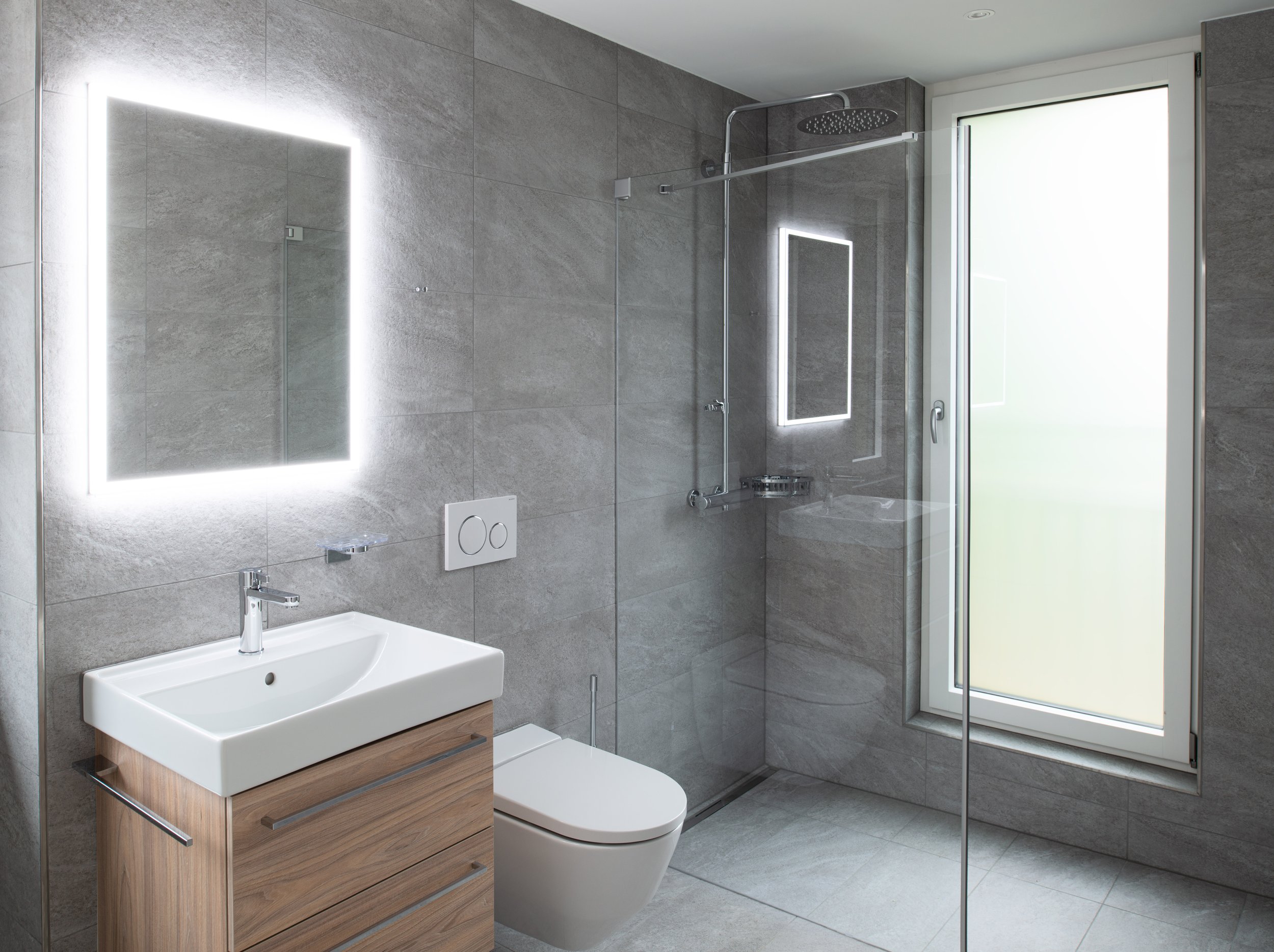 Ein modernes Badezimmer mit grauen Wandfliesen, einem rechteckigen Waschbecken auf einem Holzunterschrank, einer Toilette und einer Dusche mit Glasabtrennung. Es gibt eine beleuchtete Spiegelung über dem Waschbecken und ein Fenster mit frosted Glas.