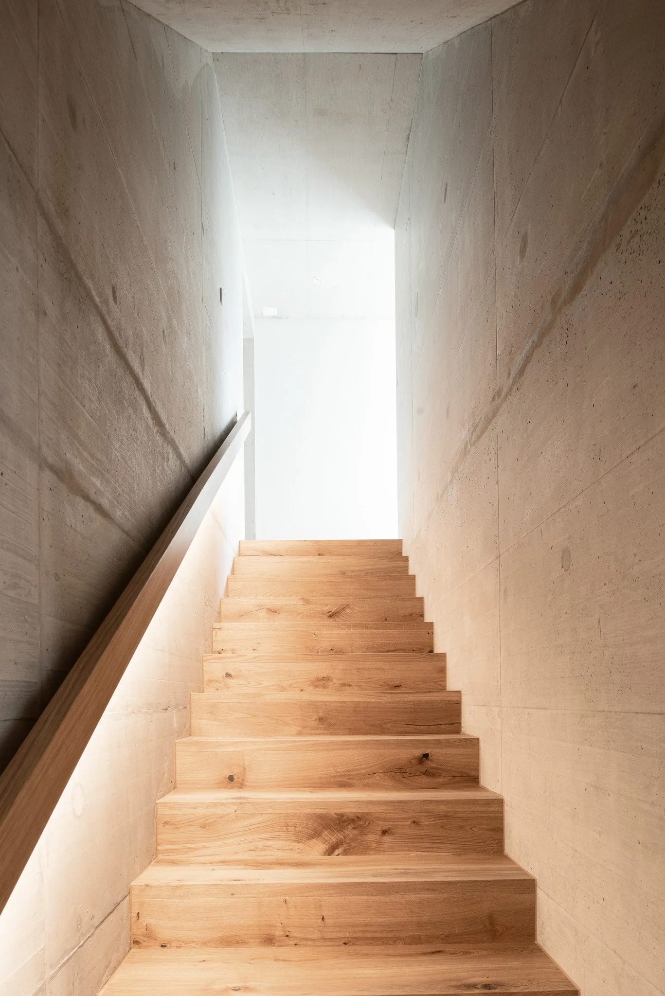 Innenansicht einer modernen Treppe aus Holz mit Betonwänden auf beiden Seiten und natürlichem Licht am oberen Ende.