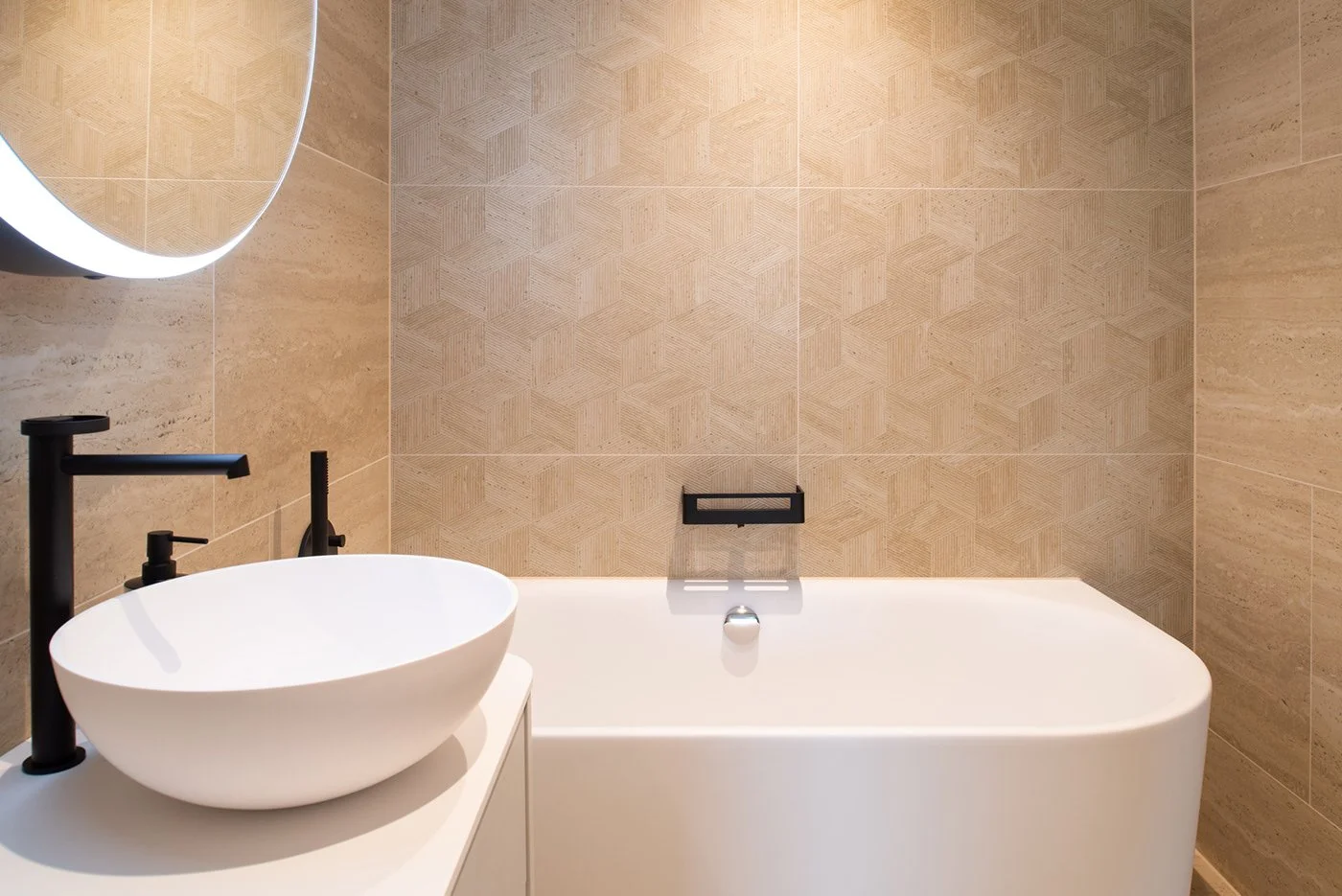 Ein modernes Badezimmer mit einem weissen Waschbecken, schwarzen Wasserhähnen und einer Badewanne vor beige Fliesen.