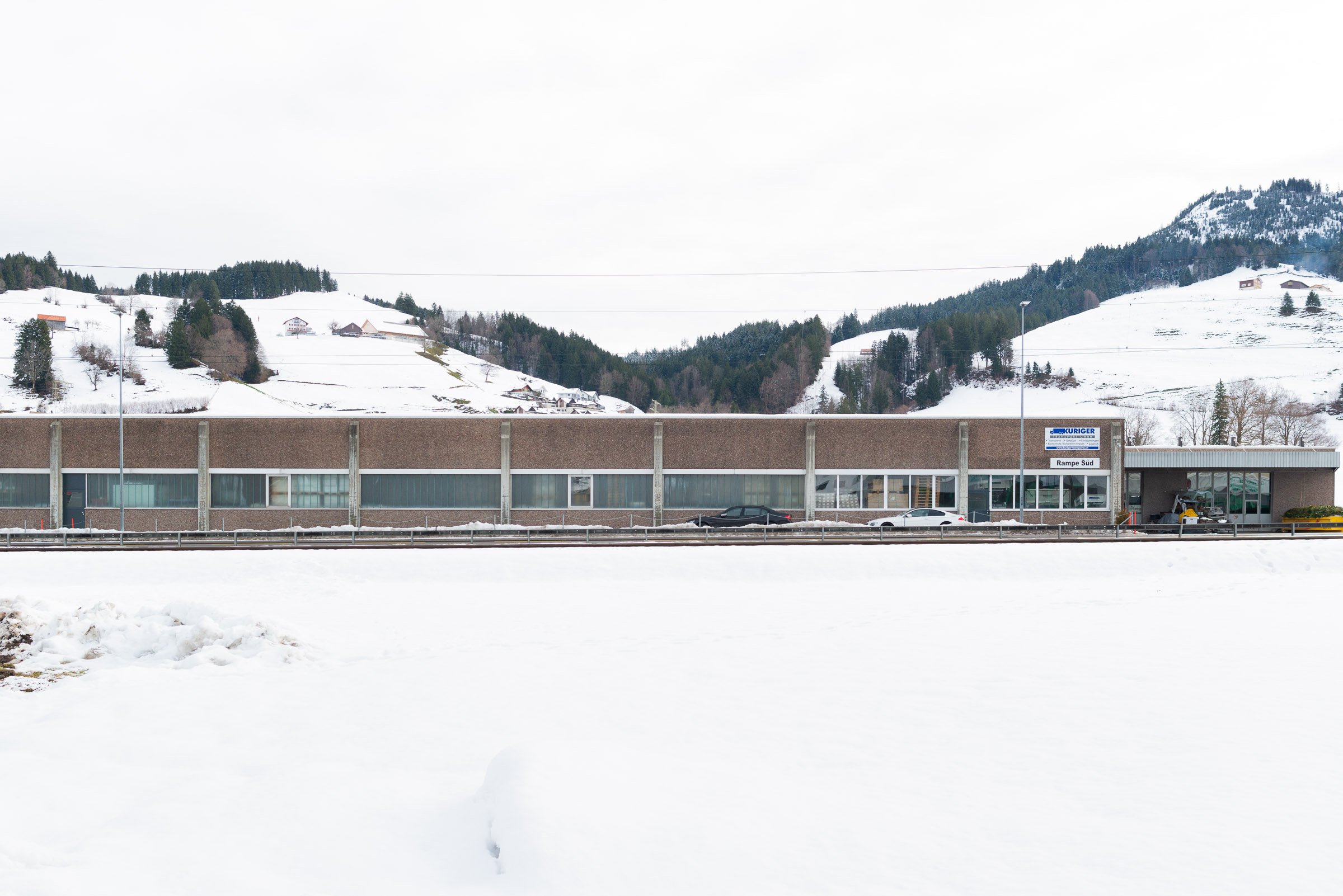 Schneebedeckter Parkplatz im Vordergrund mit einem Geschäft im Hintergrund, Berge und verschneite Hänge in der Ferne, bewölkter Himmel.