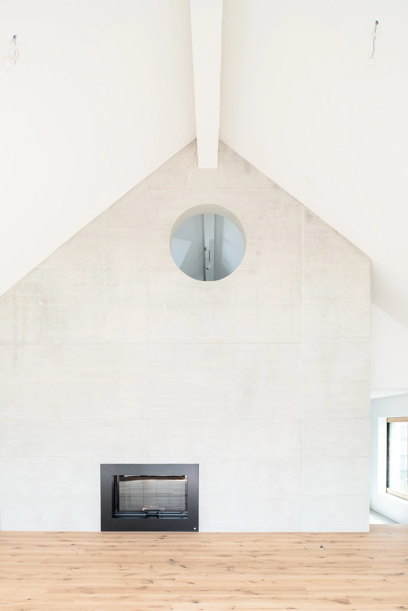 Ein modernes, minimalistisch gestaltetes Innere mit weisser Wand, Holzboden, einem schwarzen Kamin und einem runden Fenster im oberen Bereich.