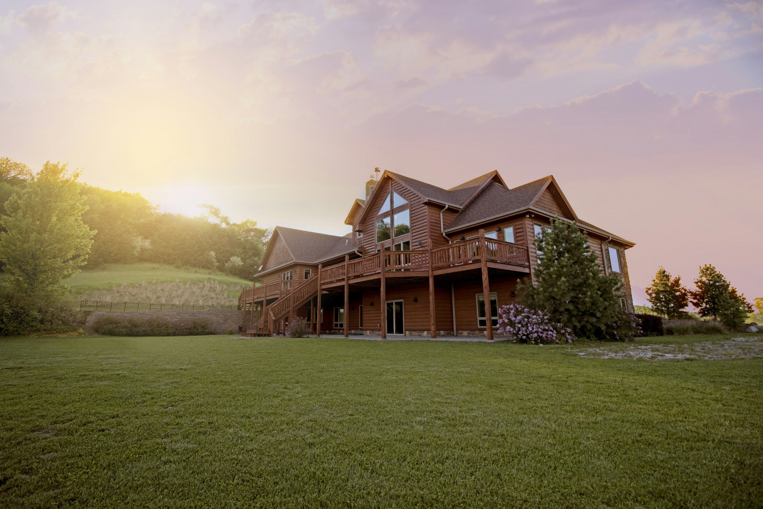 A large wooden house with a deck and multiple windows, surrounded by green grass, trees, and flowers, under a pastel-colored sky at sunset.