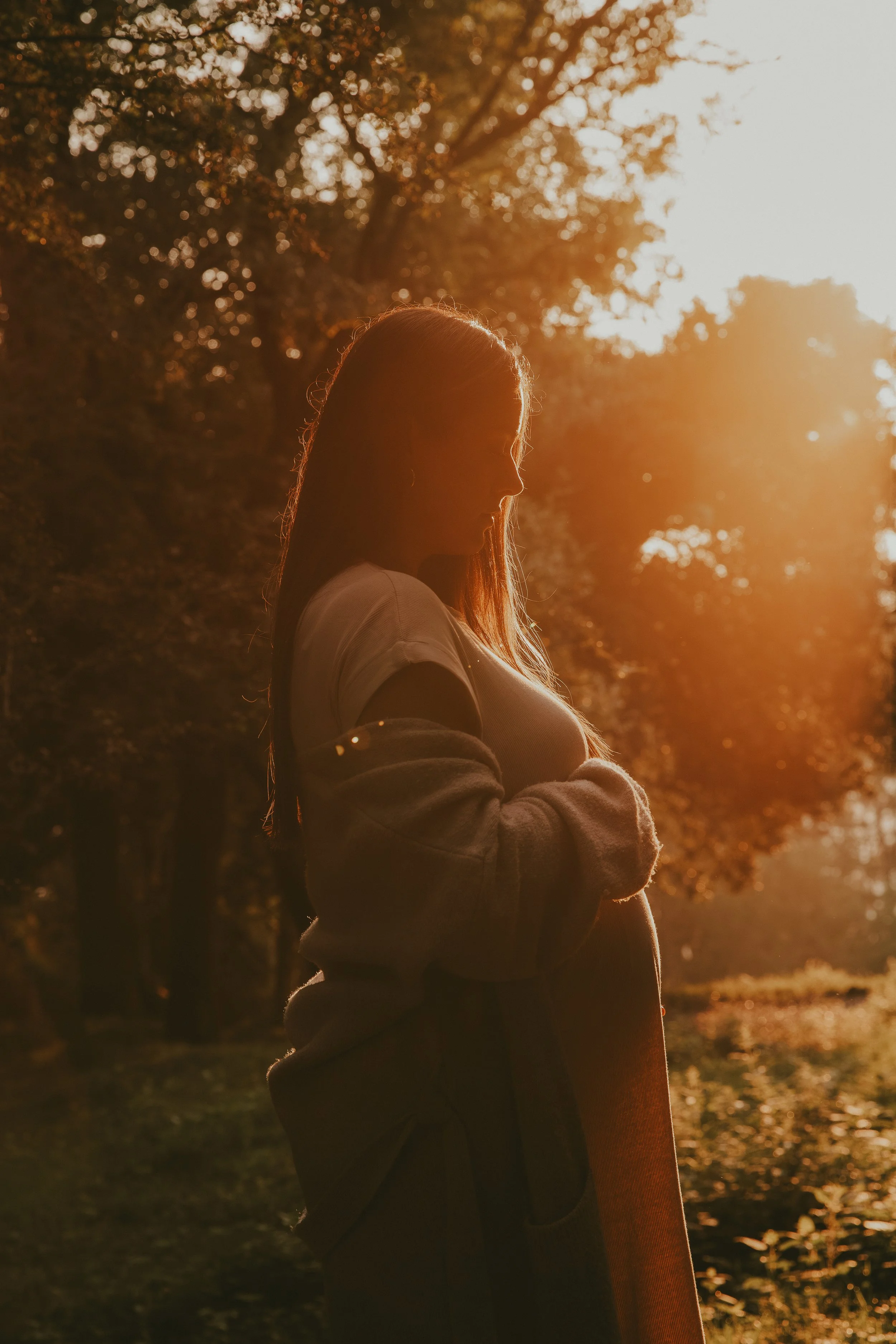 Una mujer de perfil en un entorno al aire libre durante el atardecer, con árboles y luz dorada del sol de fondo.