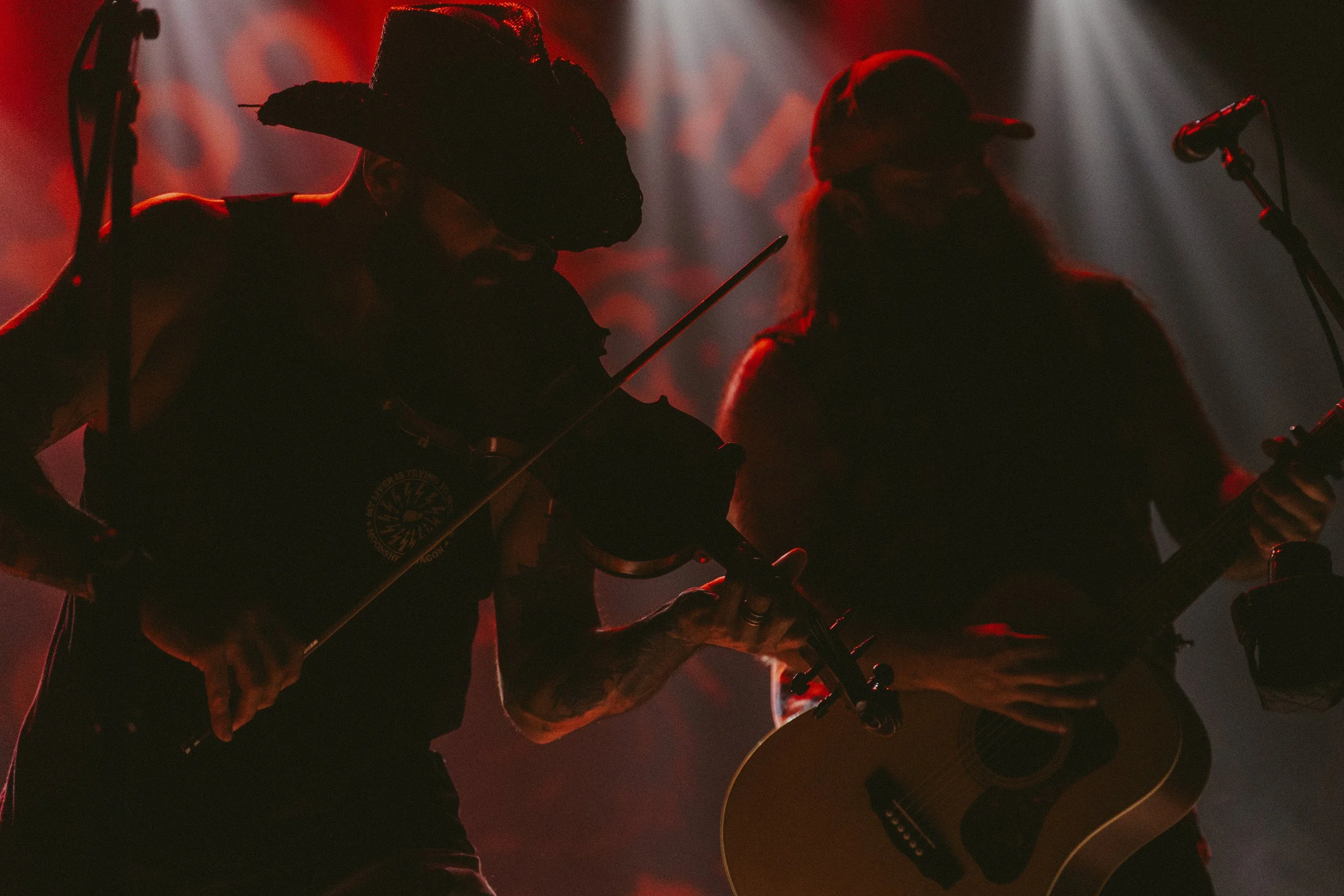 Dos músicos tocando la guitarra y el violín en un escenario con iluminación tenue y luz de fondo rojiza.