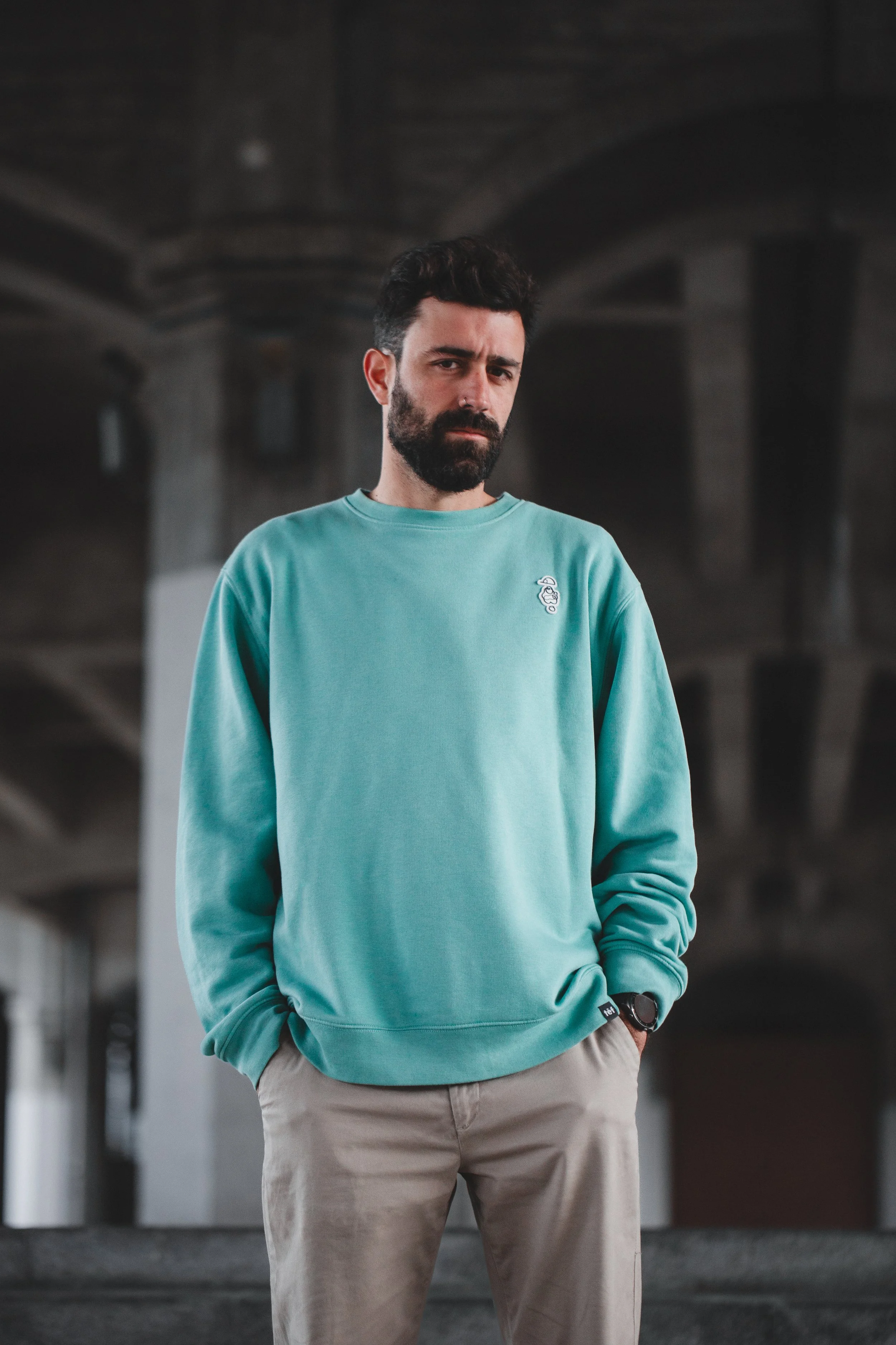 Hombre con barba en sweatshirt azul claro y pantalones beige en entorno urbano con estructura de concreto en el fondo.