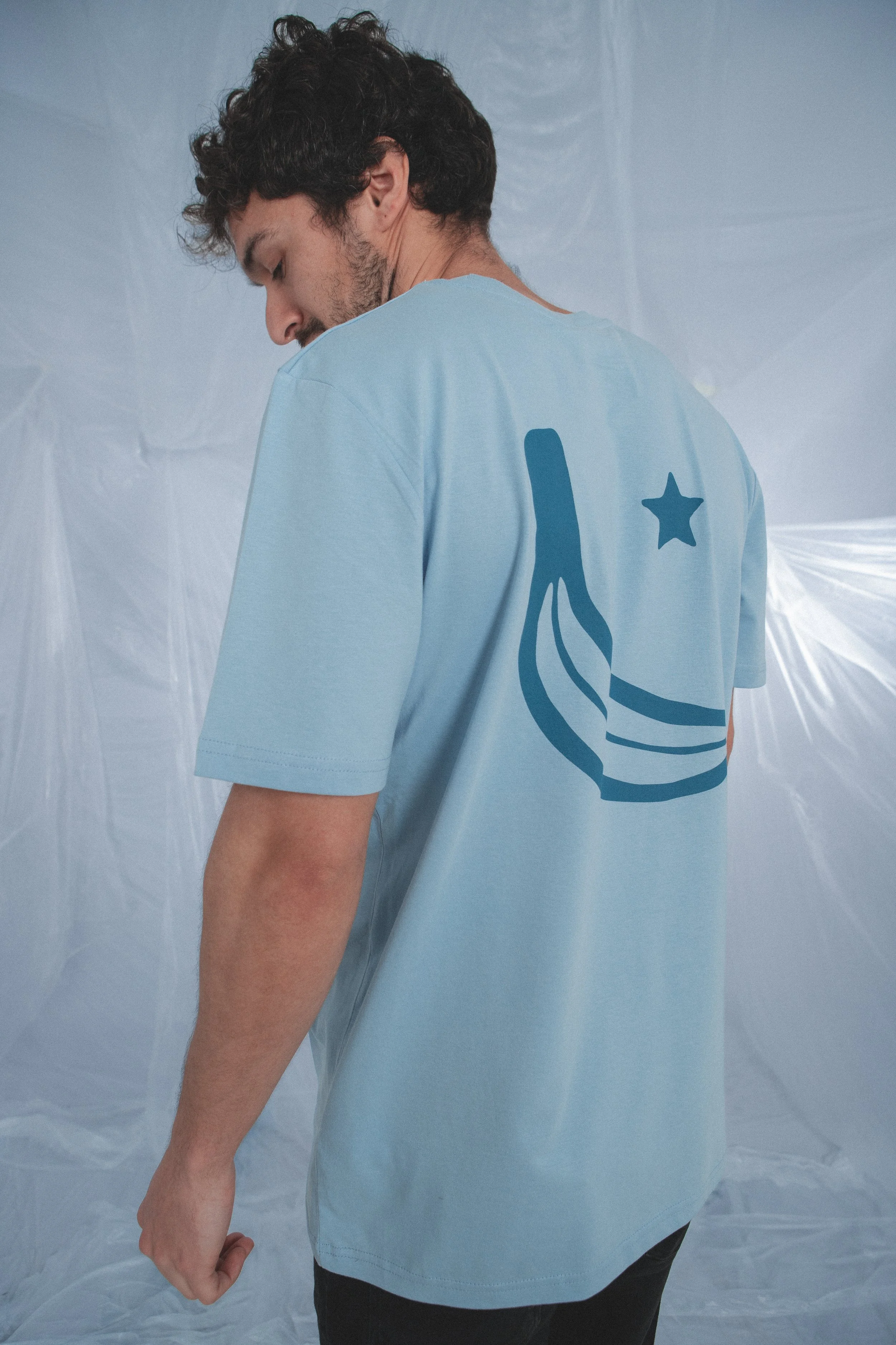 Hombre con camiseta azul que tiene un diseño en la espalda que incluye una estrella y una figura de tinta, en un fondo plástico y traslucido.