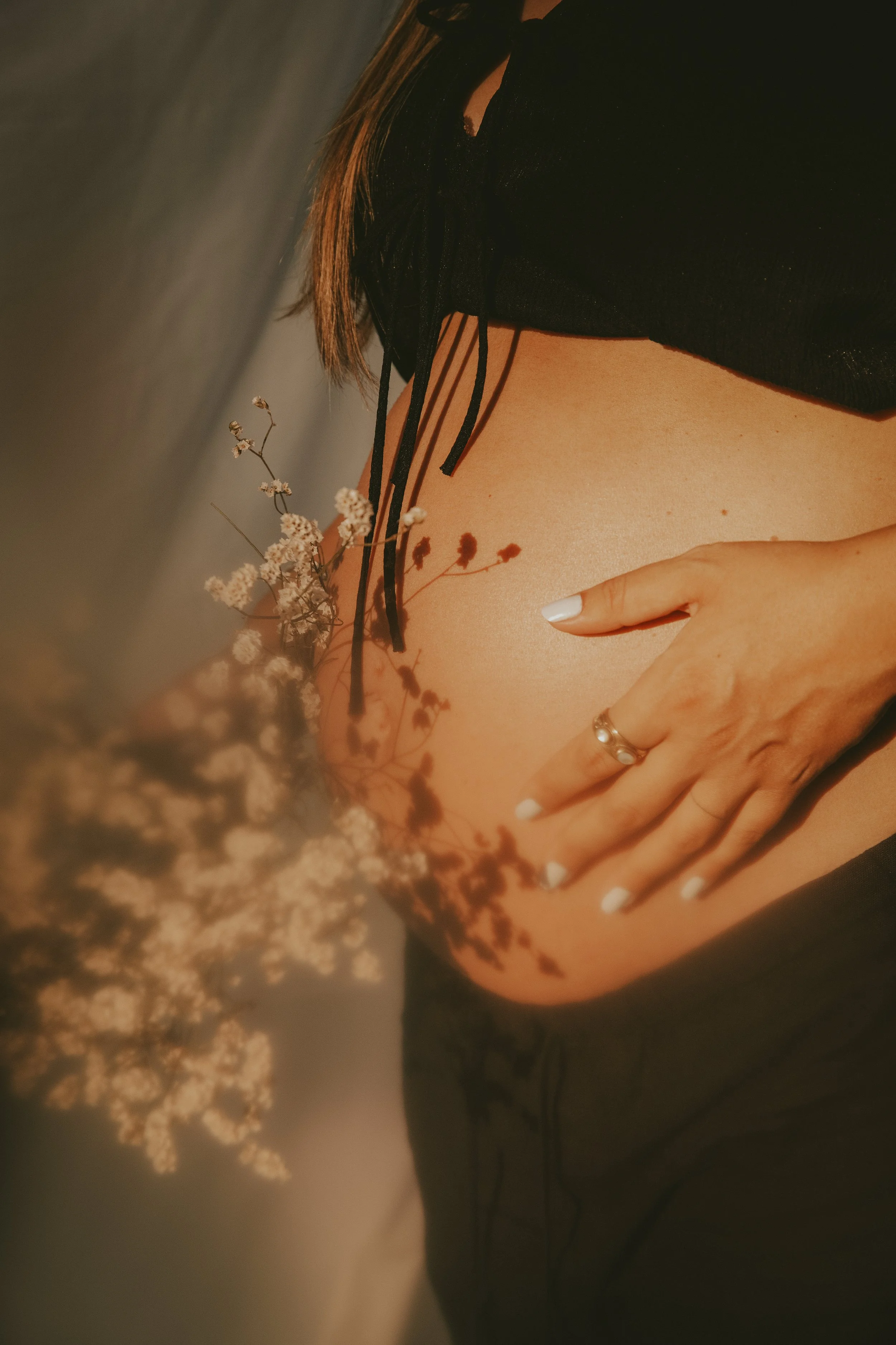 Una mujer embarazada con vestido negro, tocando su vientre y rodeada de flores secas, en una foto artística y cálida.