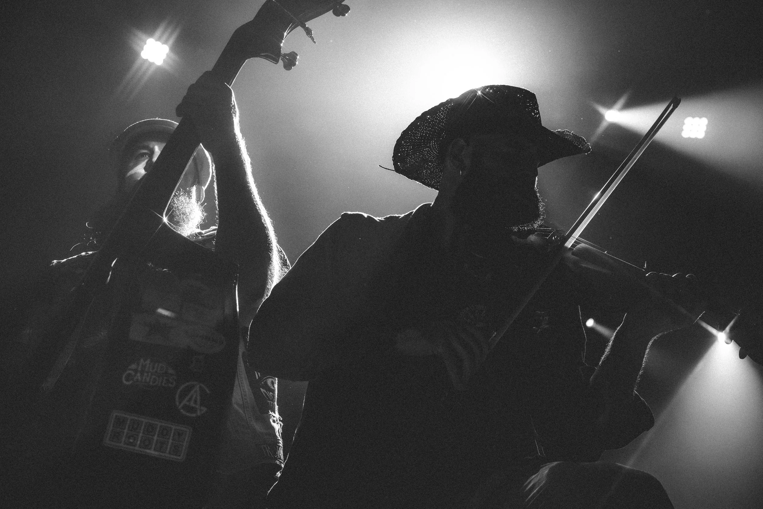 Persona tocando violín en un escenario con luces dramáticas, en silueta, usando sombrero y gafas oscuras.