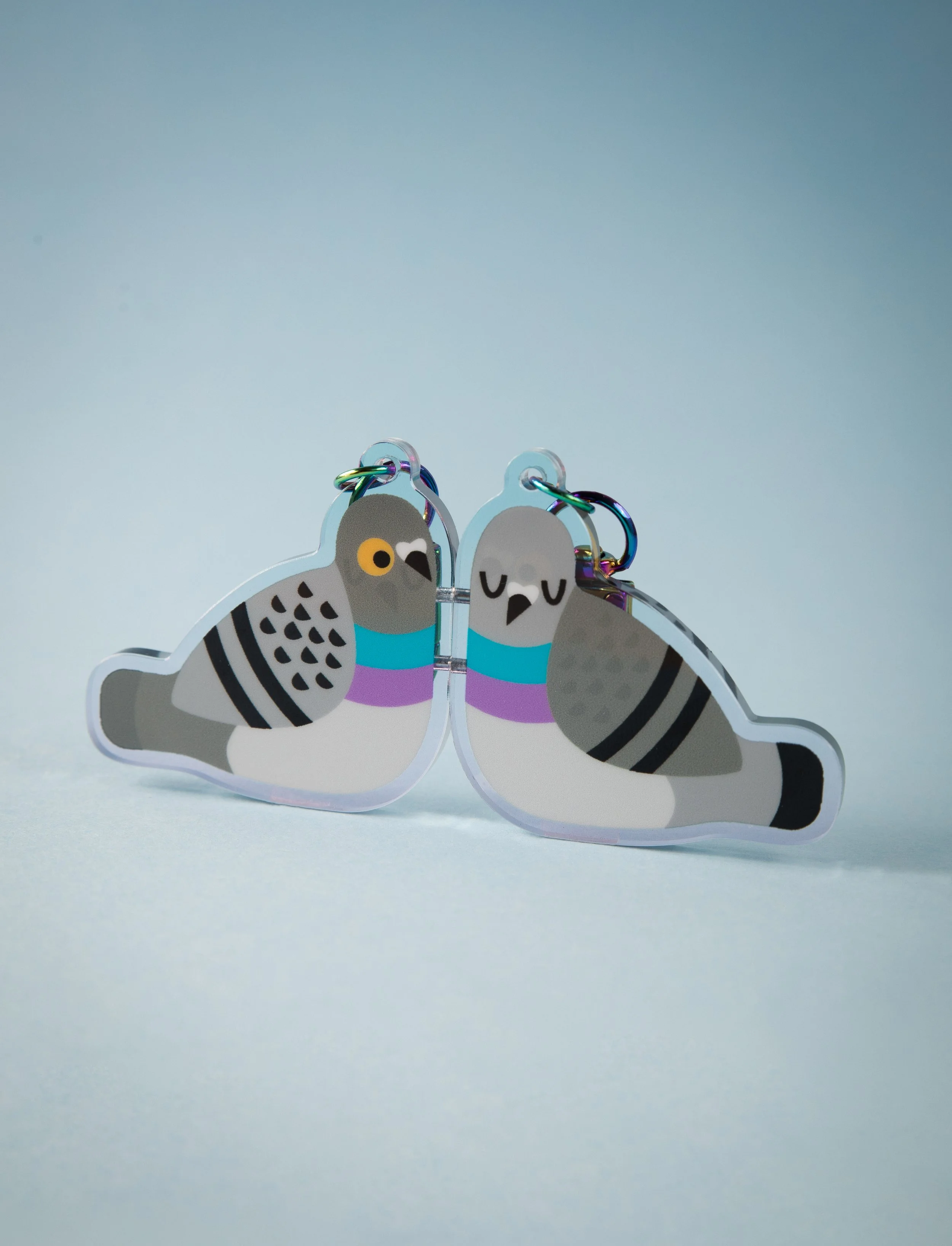 Llavero con diseño de palomas en colores pastel, una de ellas con ojos cerrados y ambas con azul y morado en sus cuellos, en un fondo de color claro.