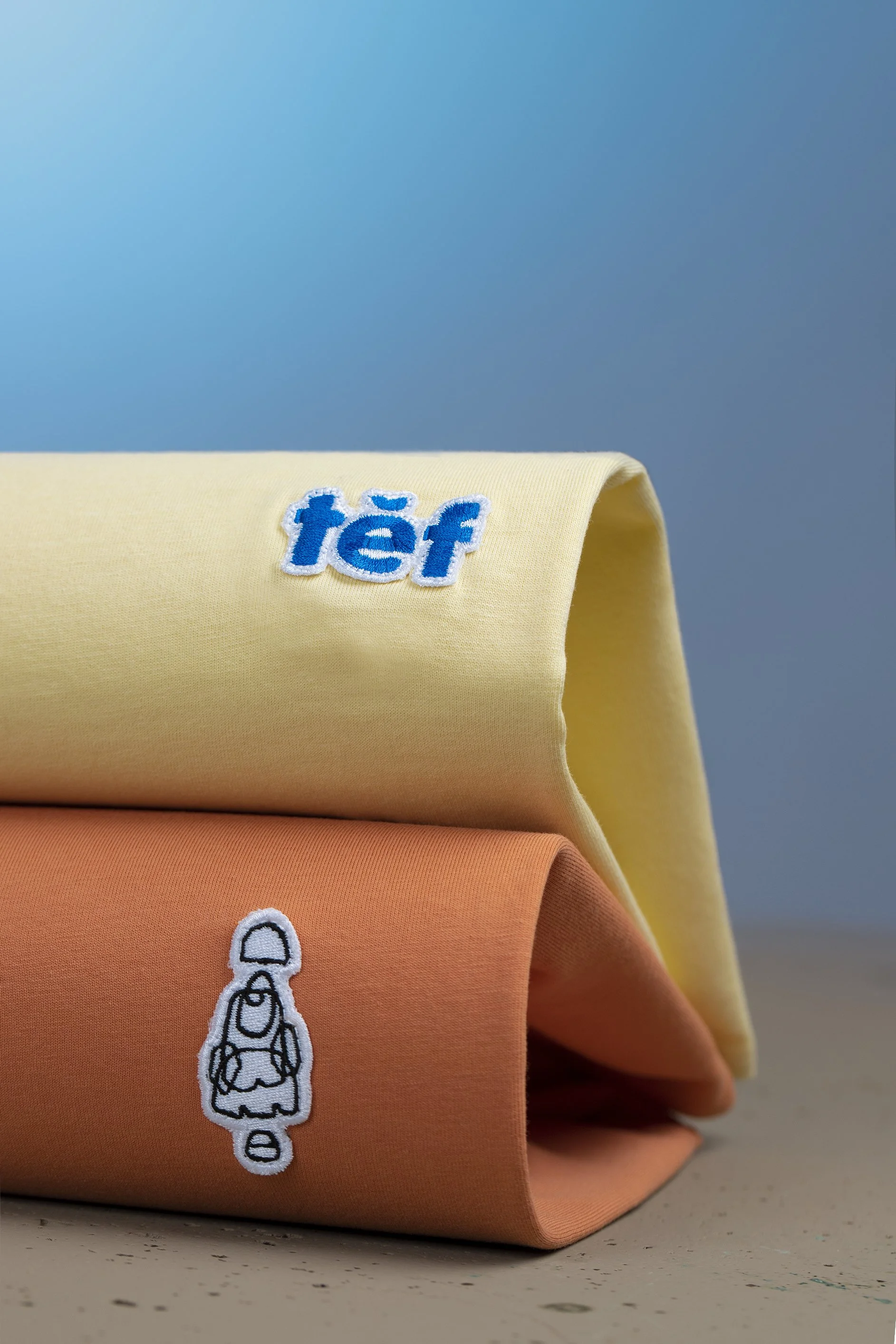 Dos camisetas dobladas, una amarilla con un logotipo azul que dice 'téf' y otra naranja con un bordado de un personaje de dibujos animados de color blanco y negro.