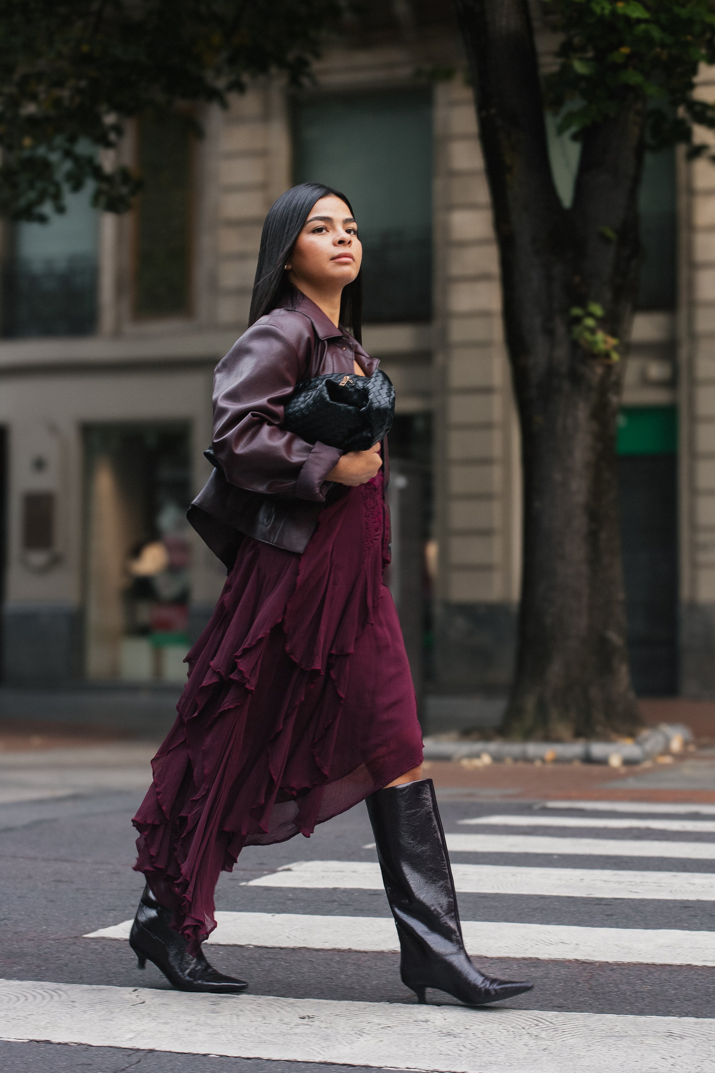 Joven mujer cruzando la calle en una ciudad con edificios de fondo, vestida con un abrigo púrpura, falda larga y botas negras de tacón alto.