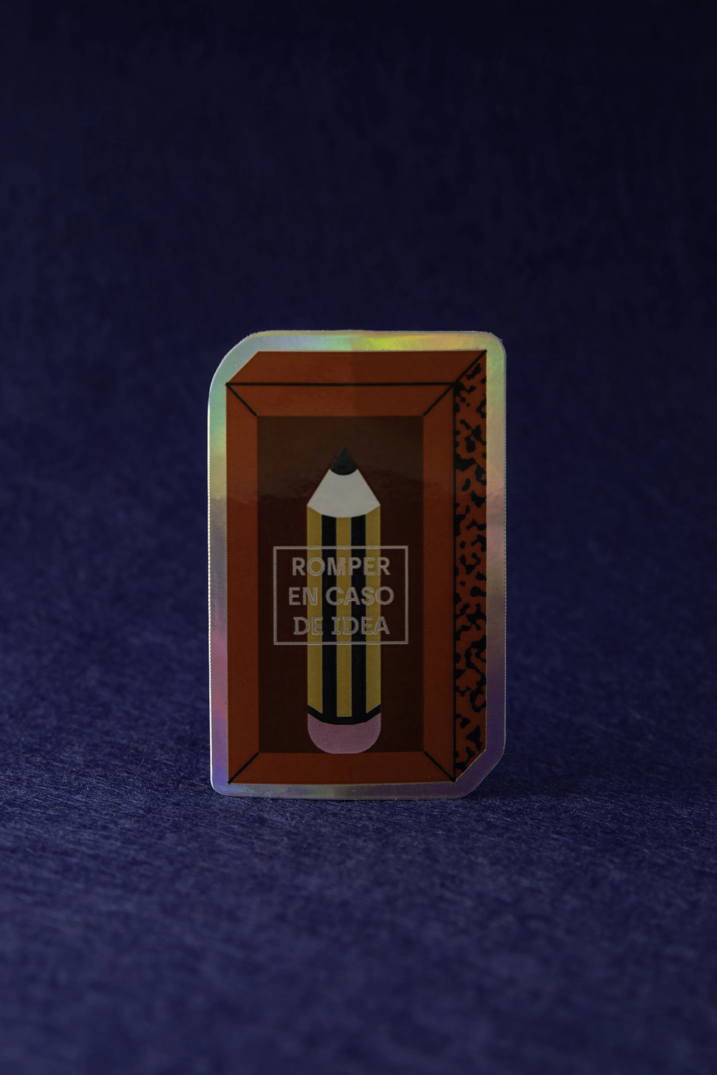 Etiqueta holográfica con una ilustración de un lápiz amarillo con rayas negras y la frase 'Romper en caso de idea' sobre fondo rojo.