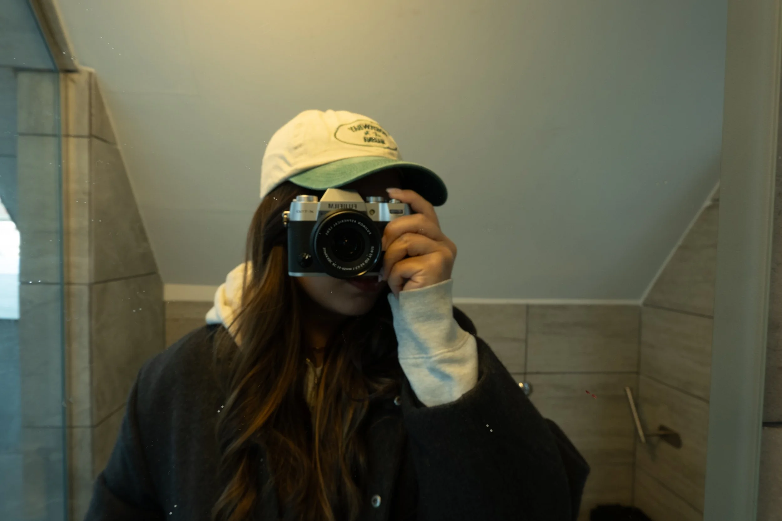Persona tomando una selfie en un espejo con una cámara, lleva una gorra y un suéter gris en un baño con paredes de cerámica beige.