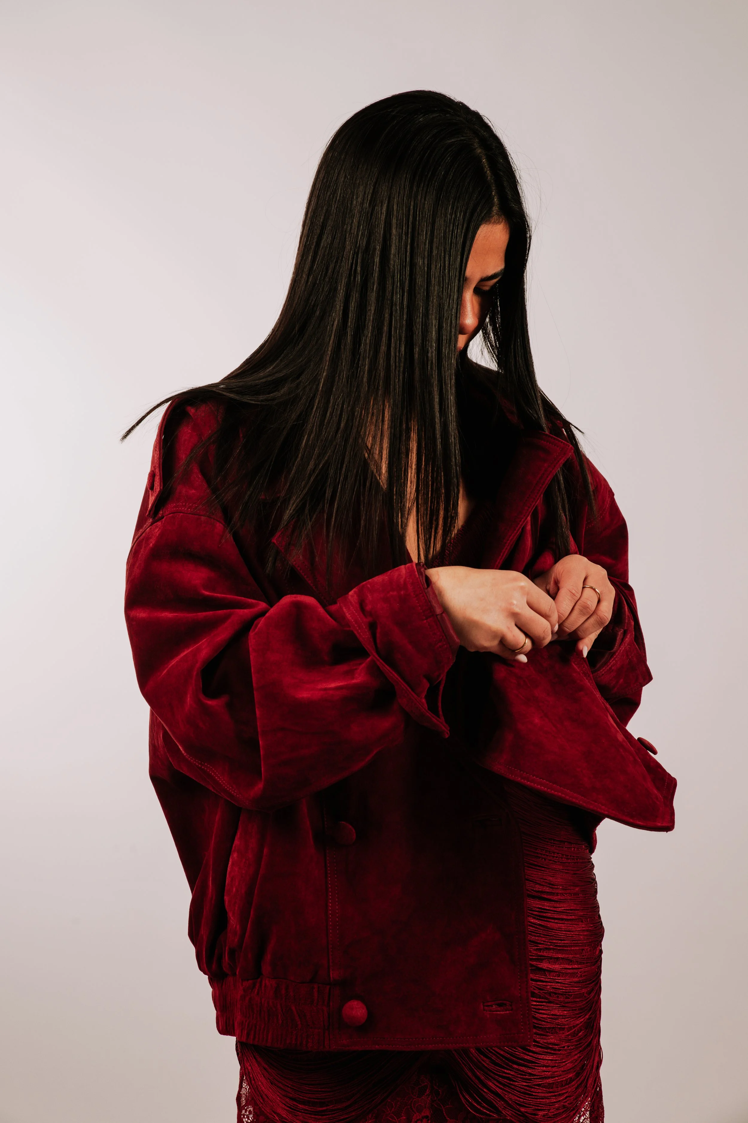 Mujer con cabello largo y oscuro, viste cazadora de terciopelo roja y falda del mismo color, ajustada y con textura arrugada, en un fondo gris claro.
