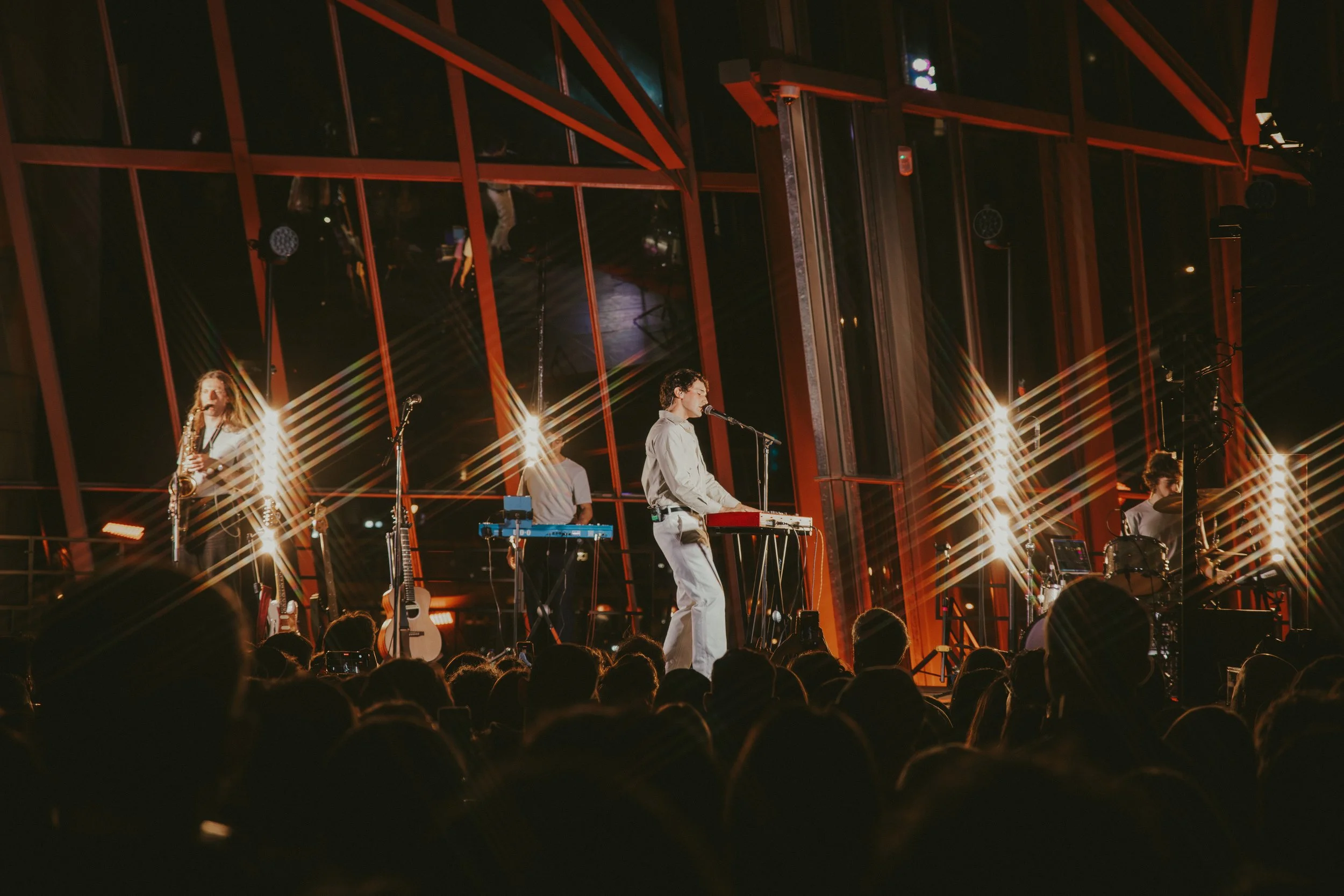 Concierto en un escenario con músicos tocando instrumentos y público en primer plano, iluminación brillante y estructura moderna con grandes ventanas de fondo.