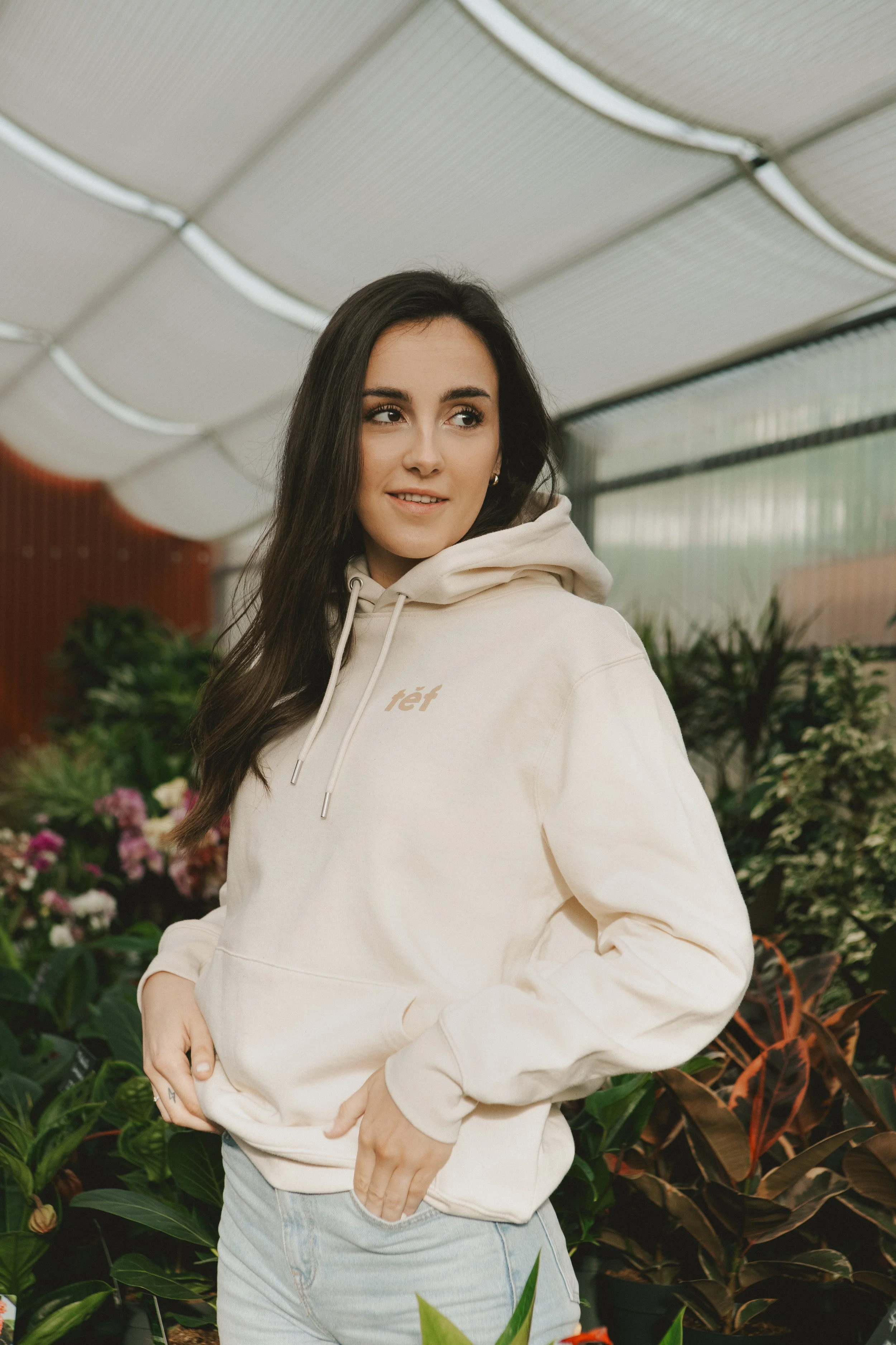 Joven mujer con sudadera beige y jeans en un invernadero rodeada de plantas y flores.