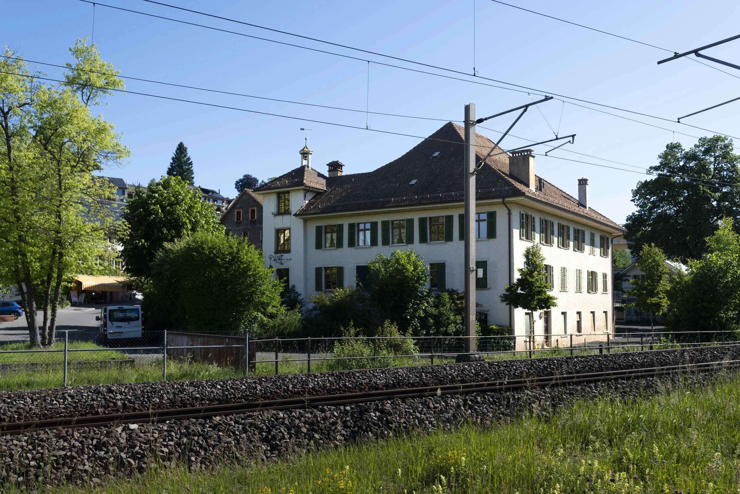A white house with green shutters surrounded by trees, with train tracks and overhead power lines in the foreground.