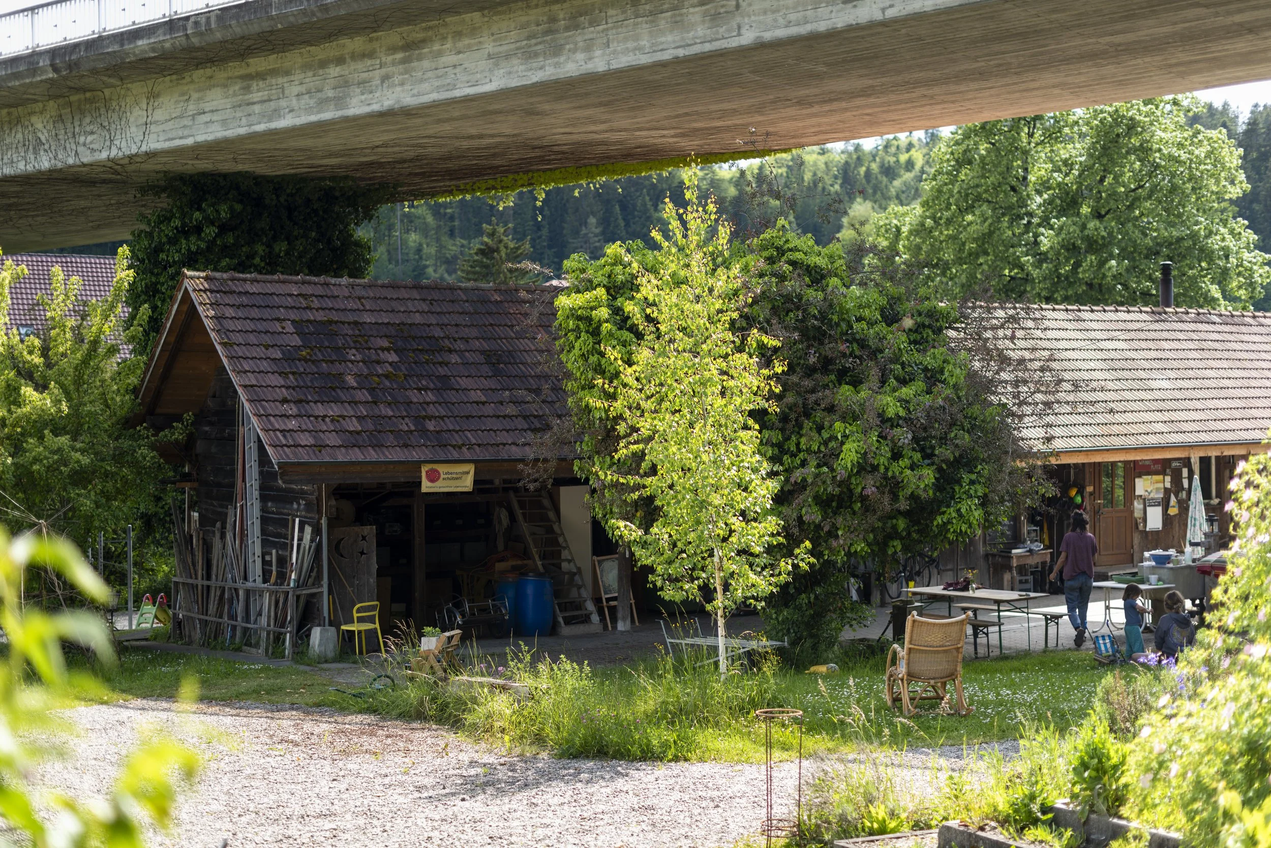 A rural outdoor scene with a large concrete bridge overhead, colorful trees, and houses with red tile roofs. There are people sitting and walking near a wooden building, with various furniture and items around, including a table with chairs and a garden area.