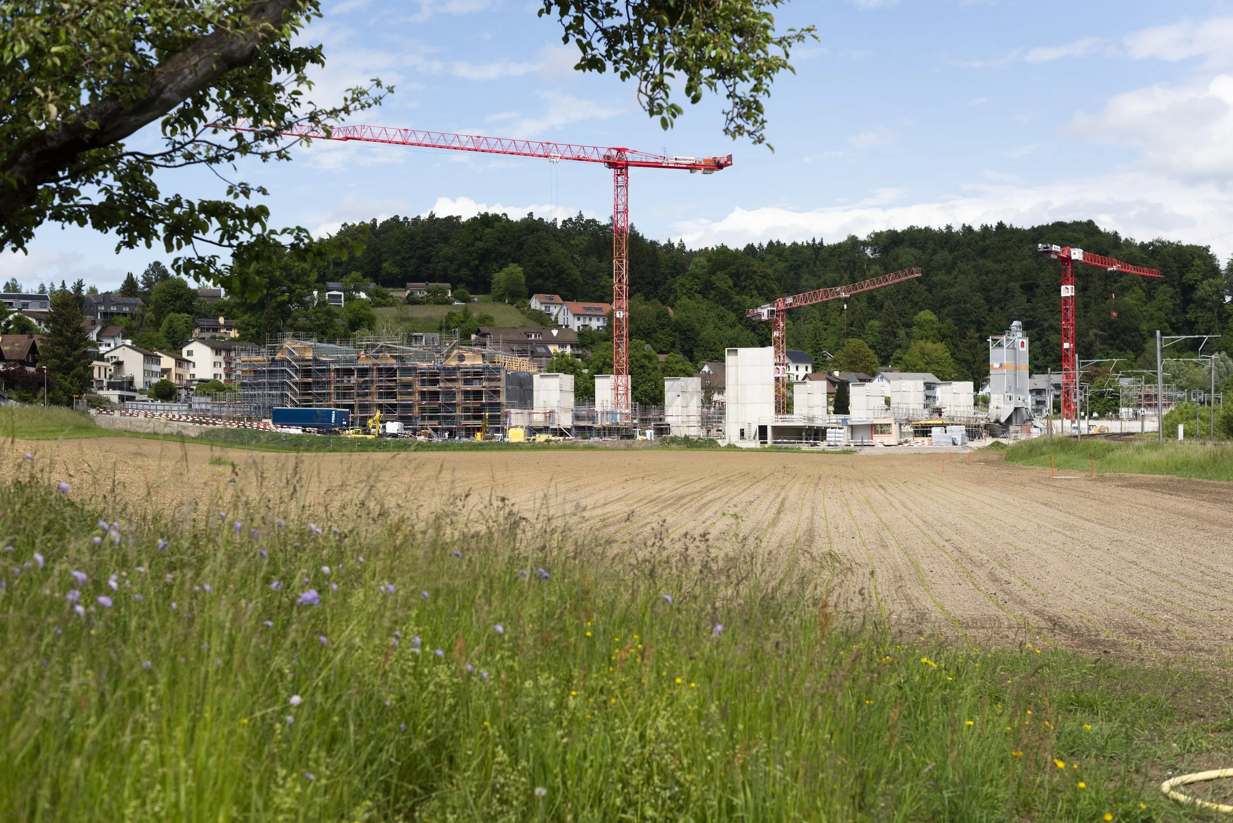 Construction site with two red tower cranes and partially built structures in a rural area with hills and trees in the background.