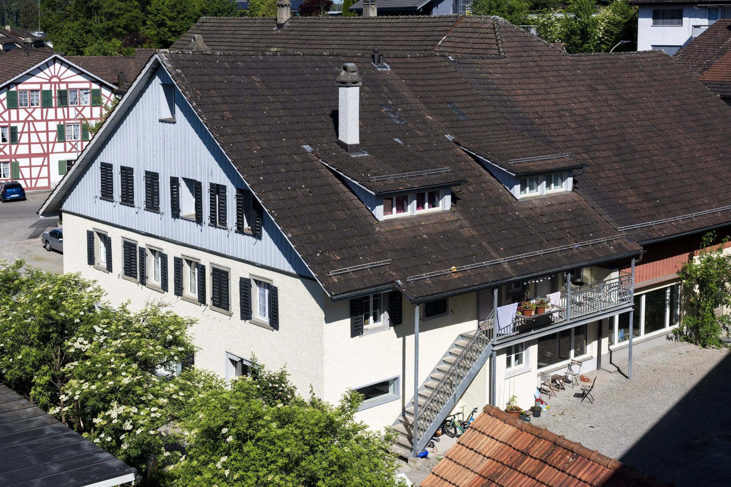 Aerial view of a residential building with a brown tiled roof, several chimney stacks, and multiple windows with black shutters. There is a small balcony with potted plants and laundry hanging outside. Green trees surround the building, and adjacent 