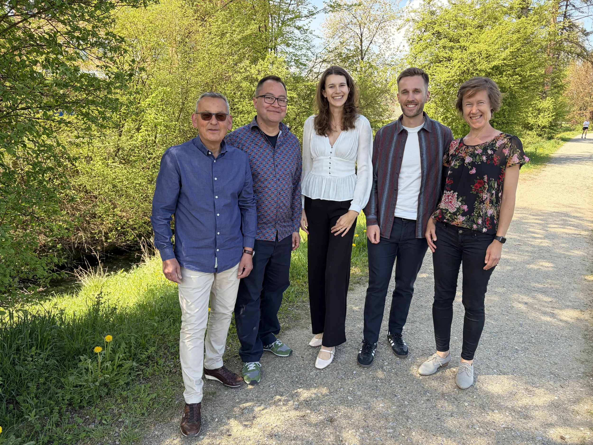 A group of five people standing together outdoors on a sunny day. They are smiling and dressed in casual and semi-formal attire. The background features green trees and a walking trail.