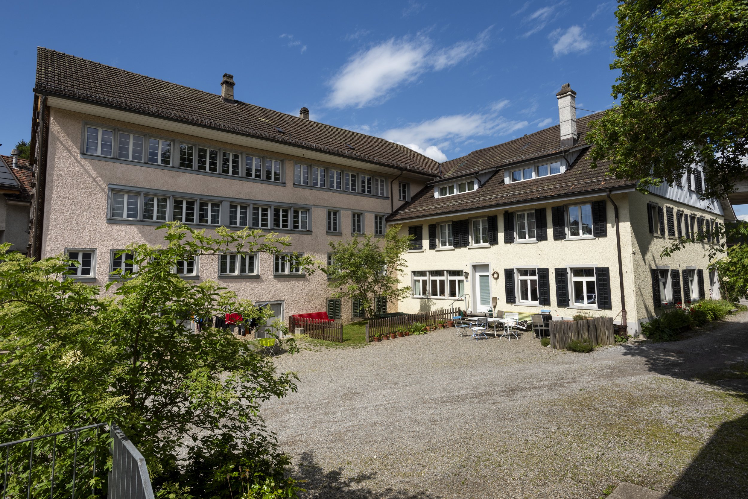 A large residential house with multiple floors, white and beige exterior walls, and a brown tiled roof. There are numerous windows on each floor and black shutters on some windows. The yard is gravel with outdoor furniture, a small garden area, and a