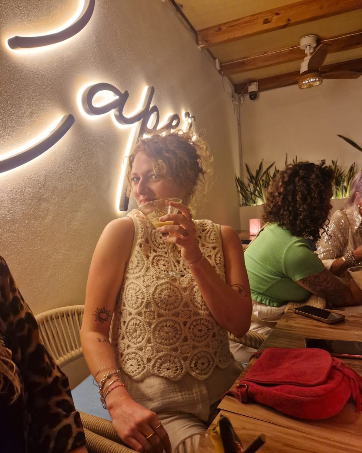 A woman with curly hair holding a cocktail inside a restaurant with a neon sign on the wall.