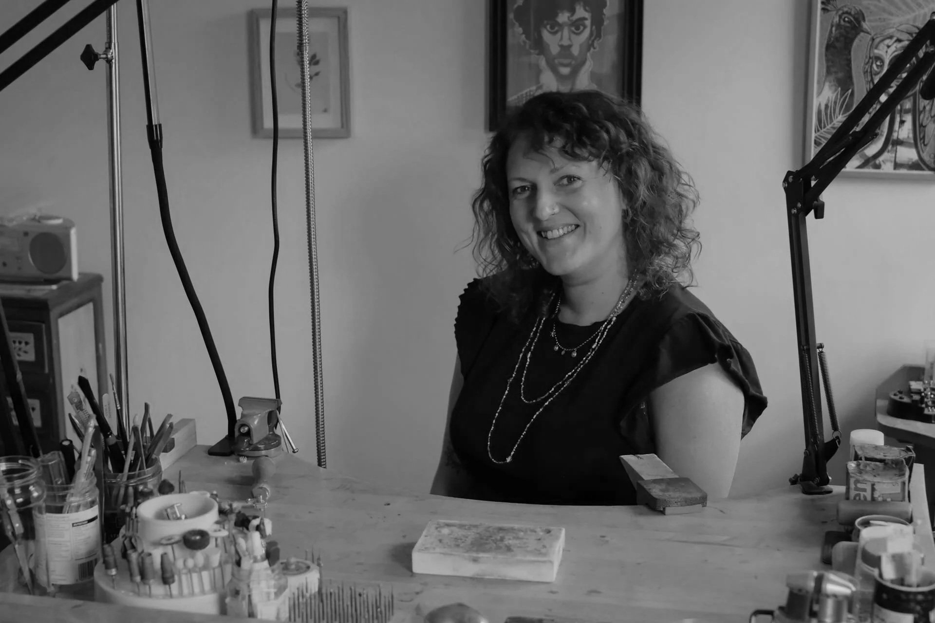 A woman smiling at a jewelry workspace with tools, jewelry pieces, and artwork behind her in black and white.