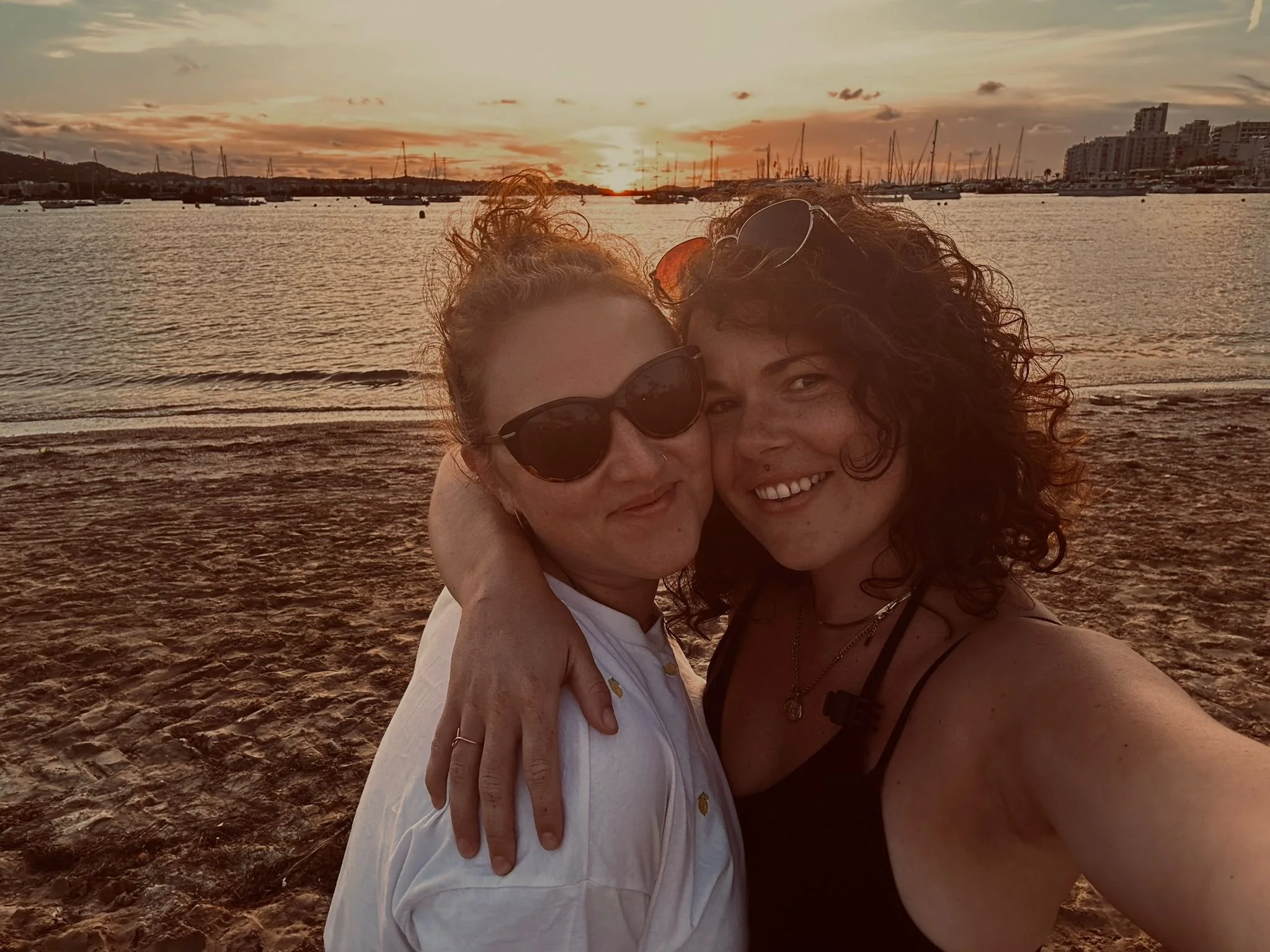 Two women pose for a selfie on a beach with sailboats and buildings in the background during sunset. One woman is wearing sunglasses and a white shirt, and the other has curly hair and a black top.