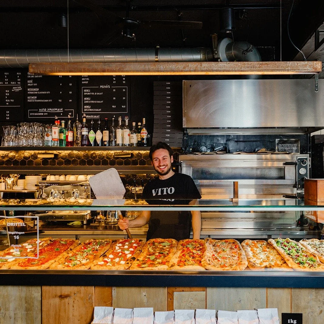 Ein Mann steht hinter einer Pizzabar-Theke mit verschiedenen Pizzen, im Hintergrund sind Flaschen, Gläser und eine Speisekarte.