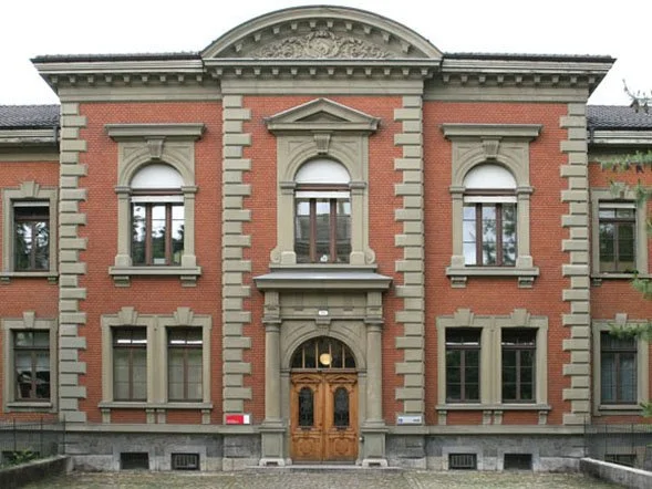 Ein mehrstöckiges altes Gebäude mit rotem Backstein und Steingewänden, große Fenster und eine Holztür im Zentrum.