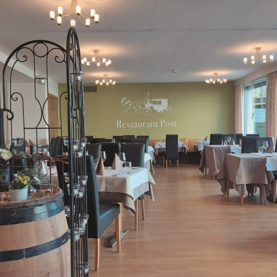 Ein leerer Speiseraum in einem Restaurant namens 'Post', mit Tischen, Stühlen, weißen Tischdecken, Geschirr und Gläsern, einer Wand mit dem Logo und dem Namen des Restaurants, großen Fenstern und modernen Deckenleuchten.