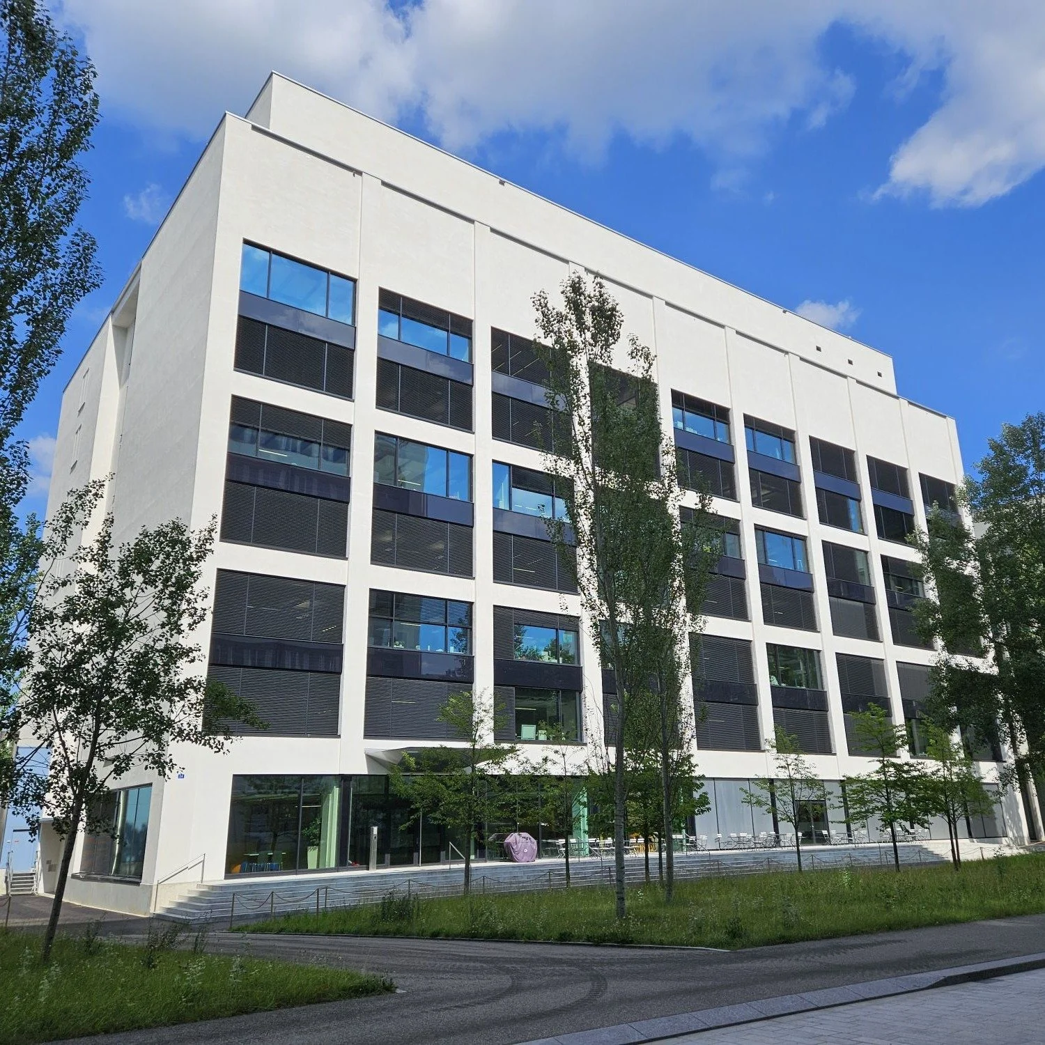 Moderne weiße Mehrgeschoss-Büro- oder Wohngebäude mit großen Fenstern, umgeben von grünen Bäumen, unter blauem Himmel mit Wolken.