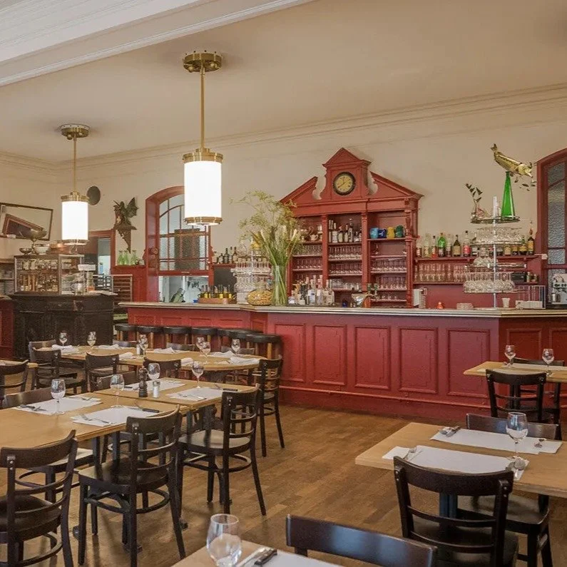 Innenraum eines Restaurants mit rotem Barbereich, Tischen mit Gläsern und Besteck, hängende Lampen, Holzboden, und dekorativen Elementen.