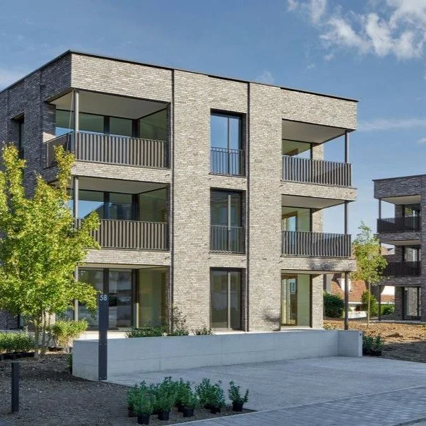 Mehrstöckiges modernes Wohngebäude mit Balkonen und Terrasse, umgeben von Pflanzen und Gehweg.