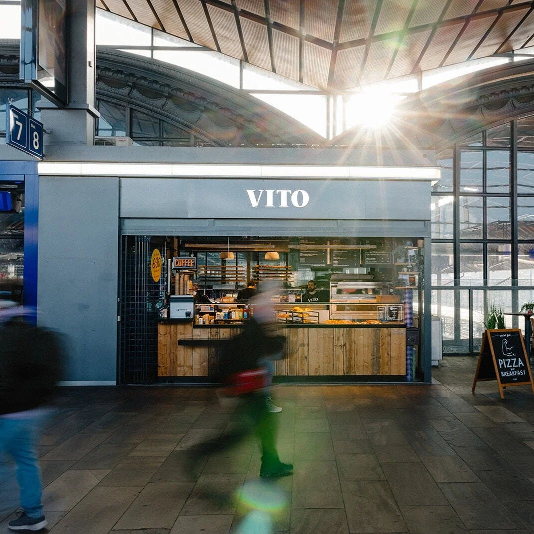 Ein Kaffeestand namens 'VITO' in einem Bahnhof. Drinnen verkaufen Mitarbeiter Kaffee und Essen, draußen steht ein kleines Pizzaschild.