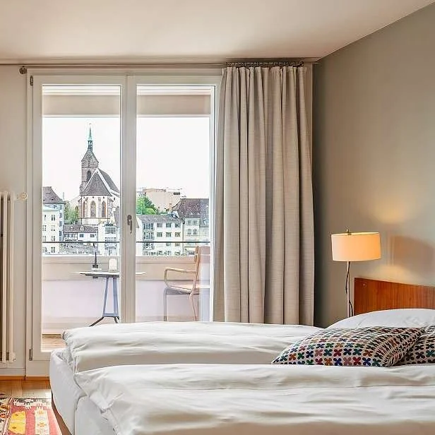 Geräumiges Hotelzimmer mit zwei Einzelbetten, Nachttischlampe, Balkon mit Blick auf eine Stadt mit Kirchturm, Vorhänge, moderner Stil.