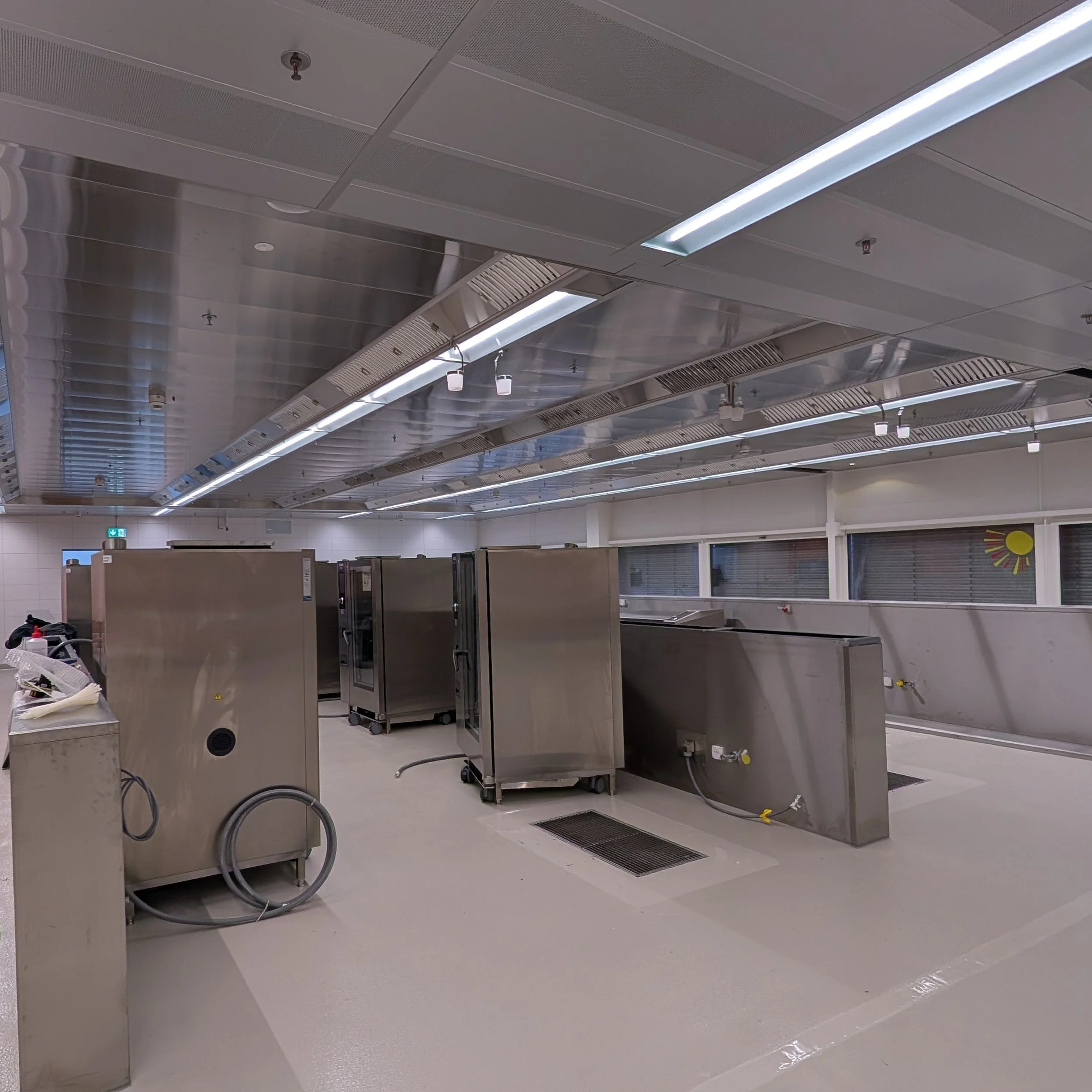 Leere Edelstahlküche mit mehreren Waschbecken, Schränken, ventilatoren und Deckenbeleuchtung.