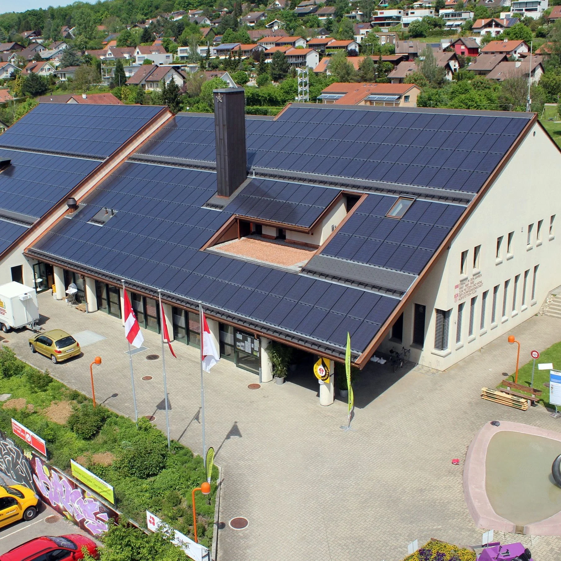 Aufnahmen eines Gebäudes mit Solarpanelen auf dem Dach, umgeben von einem Parkplatz mit Fahrzeugen und Flaggen. Im Hintergrund eine Wohnsiedlung in hügeligem Gelände mit viel Grün.