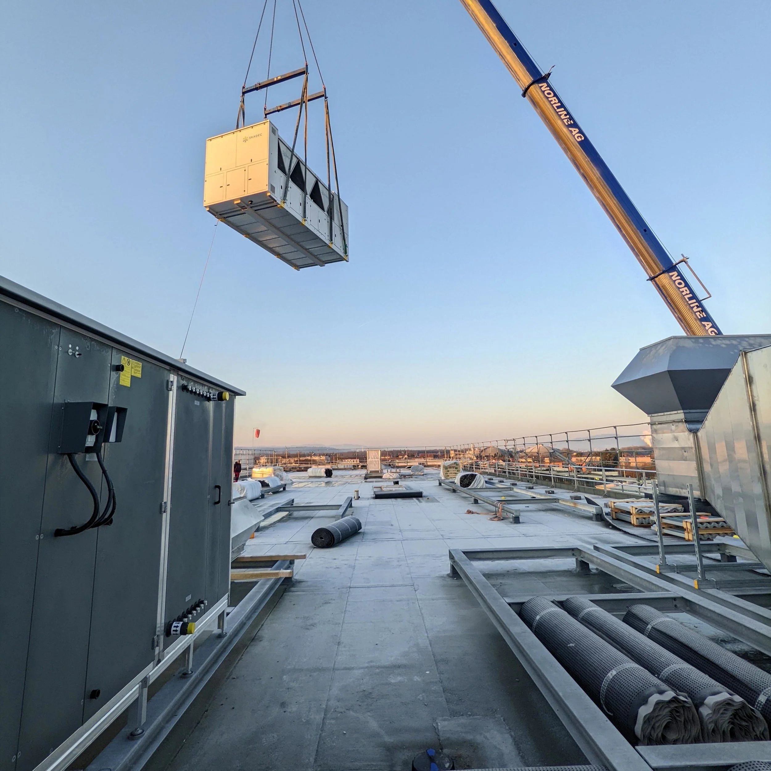 Auf einem Flachdach wird eine große Klimaanlage mithilfe eines Krans in die Luft gehoben. Im Vordergrund sind Kabel und Rohrleitungen sichtbar, im Hintergrund das Stadtbild bei Sonnenuntergang.