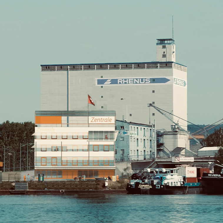 Ein Industriegebäude mit den Schriftzügen Rhenus und Zentrale, neben einem Fluss mit anlegenden Booten, an einem klaren Tag.