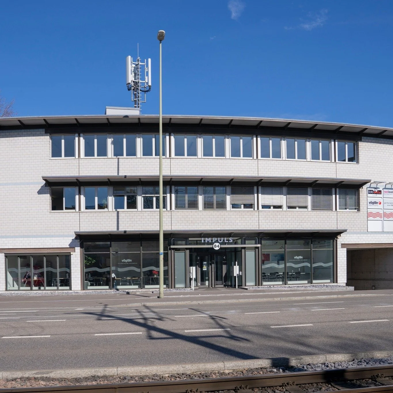 Mehrstöckiges im Bürogebäude befindliches Gebäude mit großem Eingang und Firmenlogo, vor dem Straßenbereich mit Bahnlinie.
