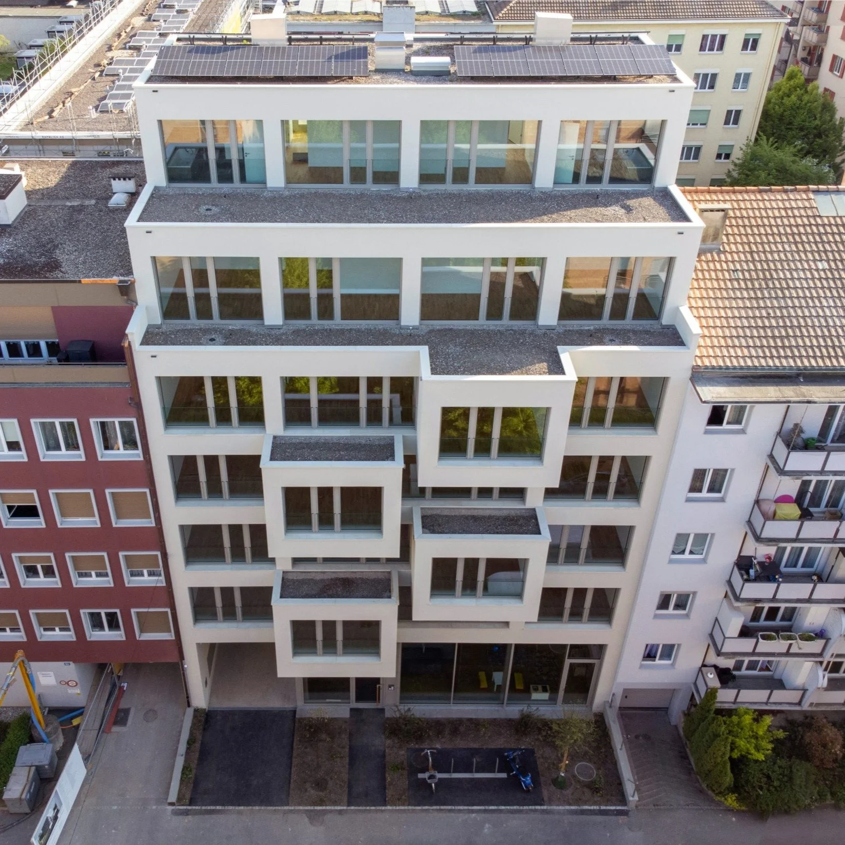 Hochhaus mit weißer Fassade und großen Fensterflächen, darunter ein Innenhof, auf dem sich Kinder mit Spielzeug befinden. Solarpanele sind auf dem Dach des Gebäudes installiert.